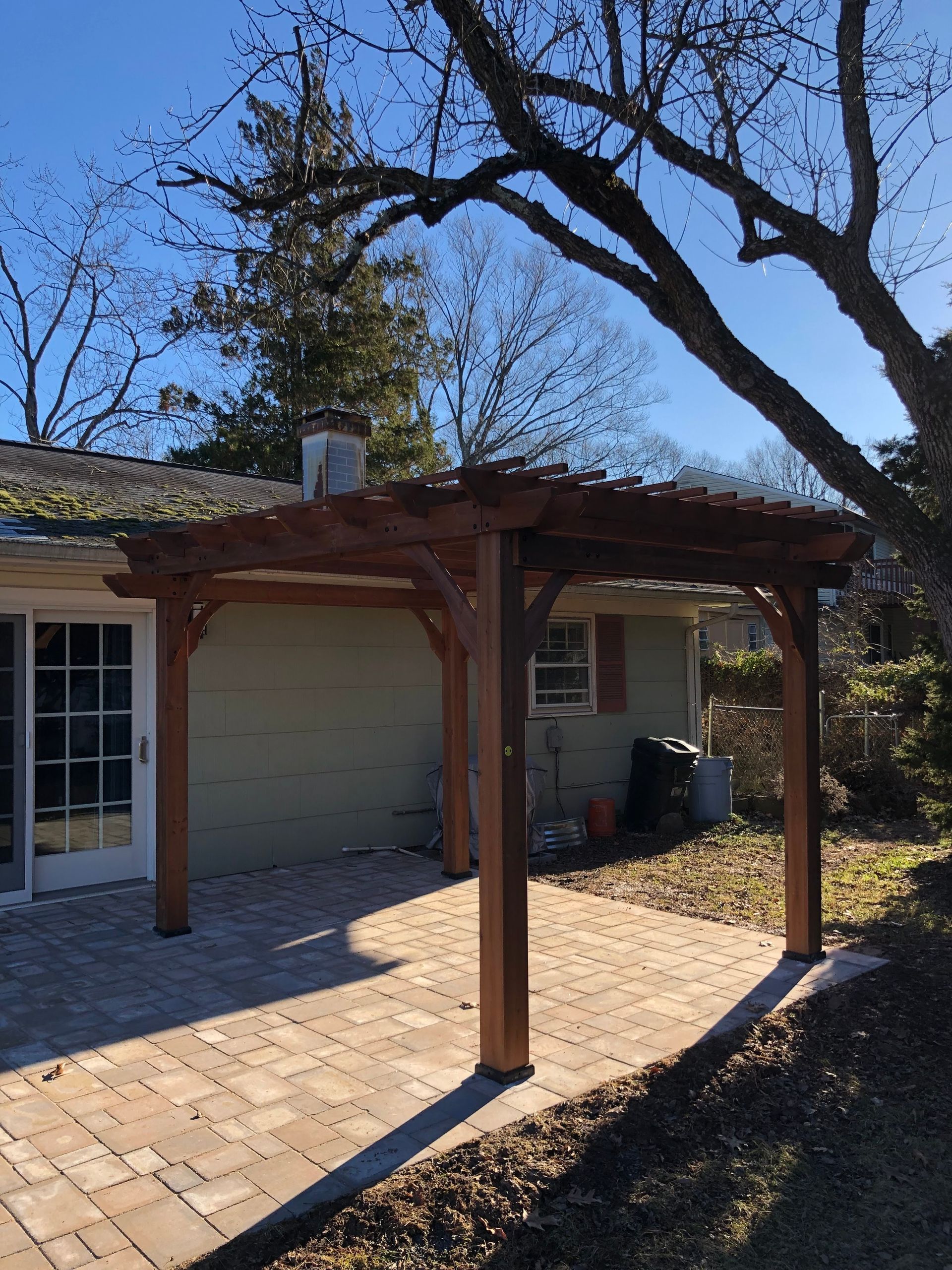 Wooden pergola beside a house with a paved patio and bare tree branches overhead