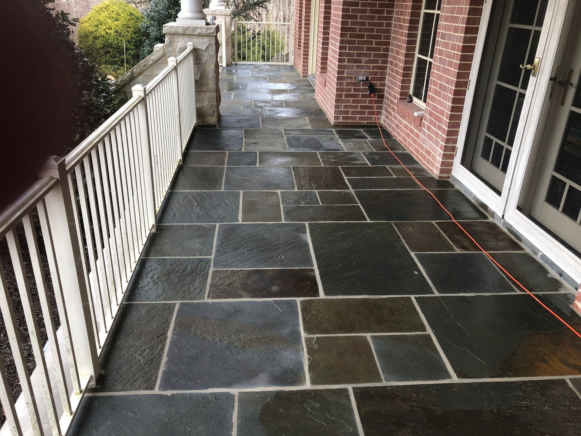 Stone patio walkway beside a brick house, with white railing and dark slate tiles.