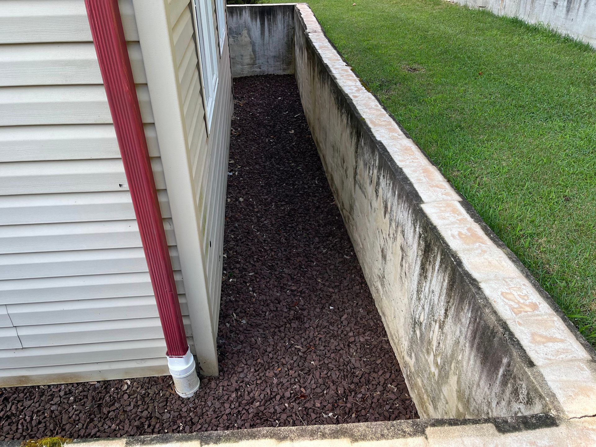 Narrow gravel strip beside a house foundation and grassy lawn, bordered by concrete walls and a downspout.