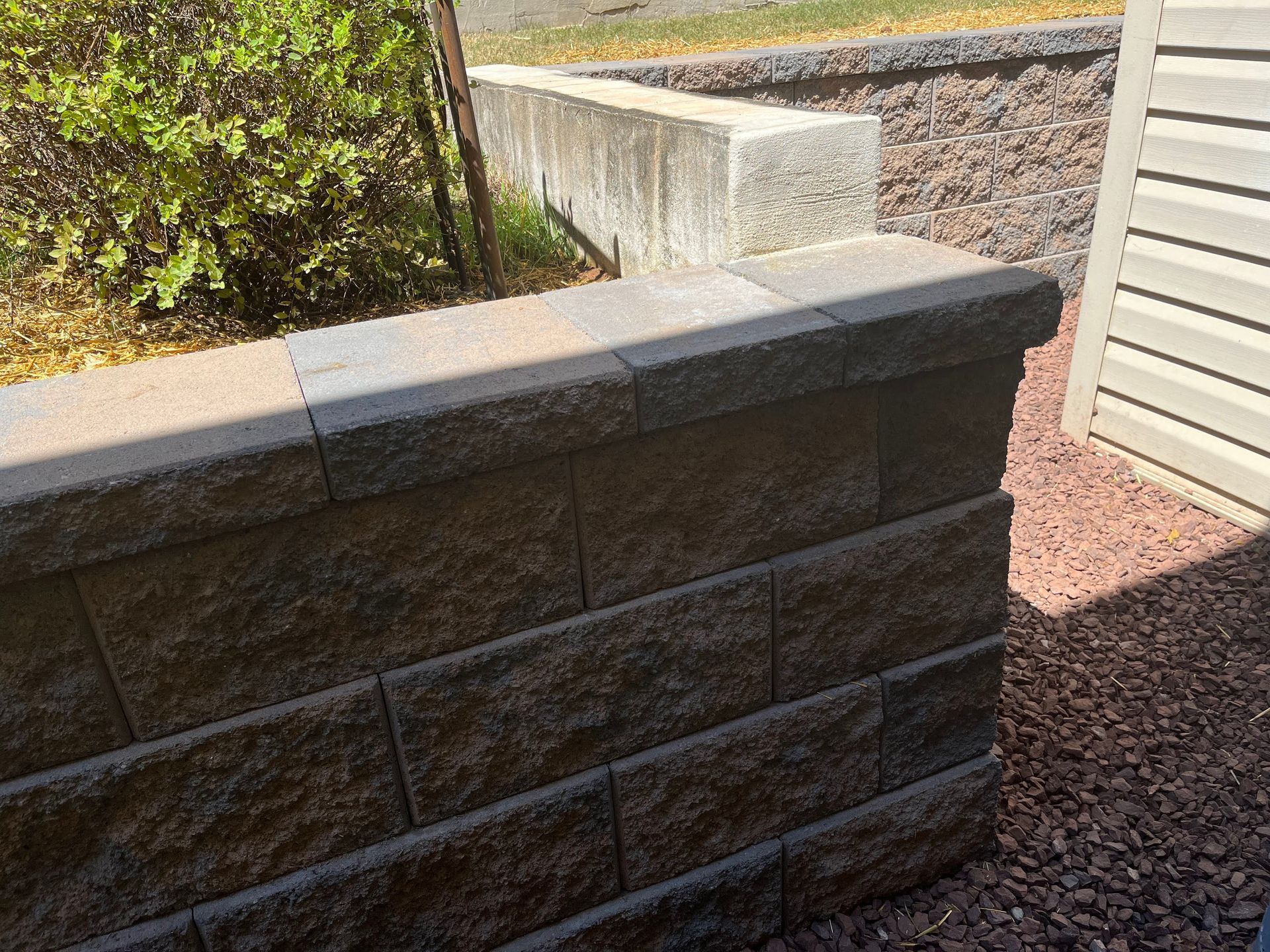 Stone retaining wall with a light gray cap along a gravel path beside a house and shrubs.
