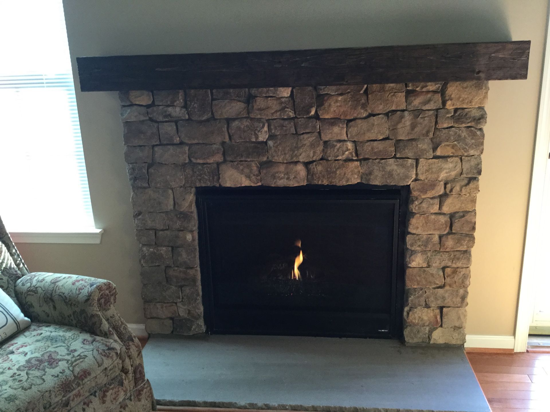 Stone fireplace with dark mantel and a small flame in the firebox, next to a window and floral armchair.