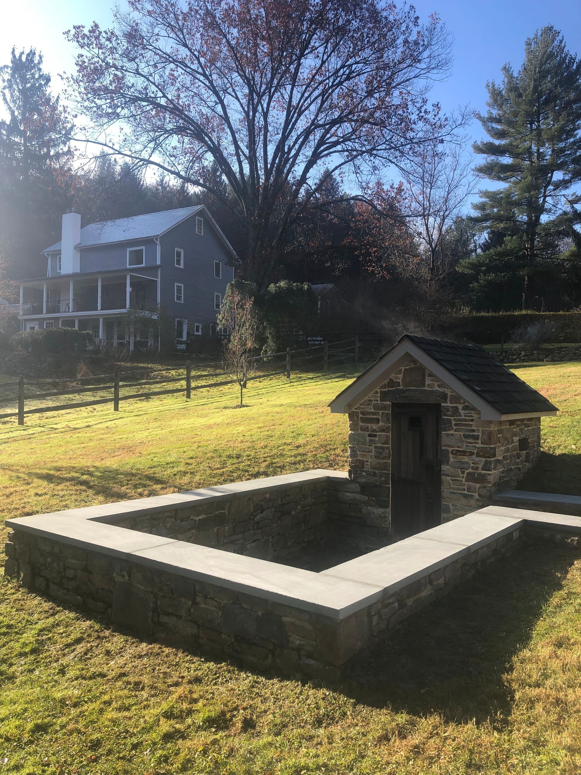 Stone well in a grassy yard with a gray house and trees in the background.