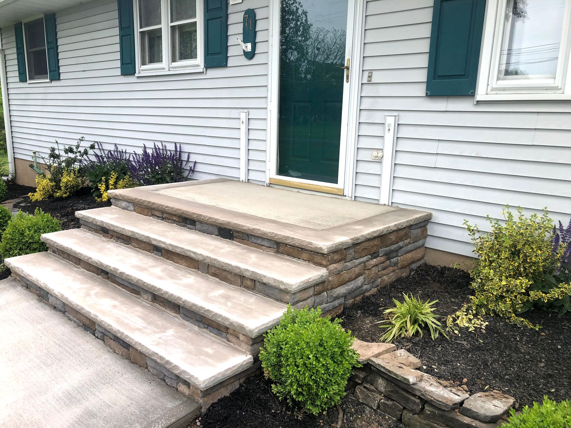 Front steps and porch of a white house with teal door and shutters, bordered by small shrubs and flowers