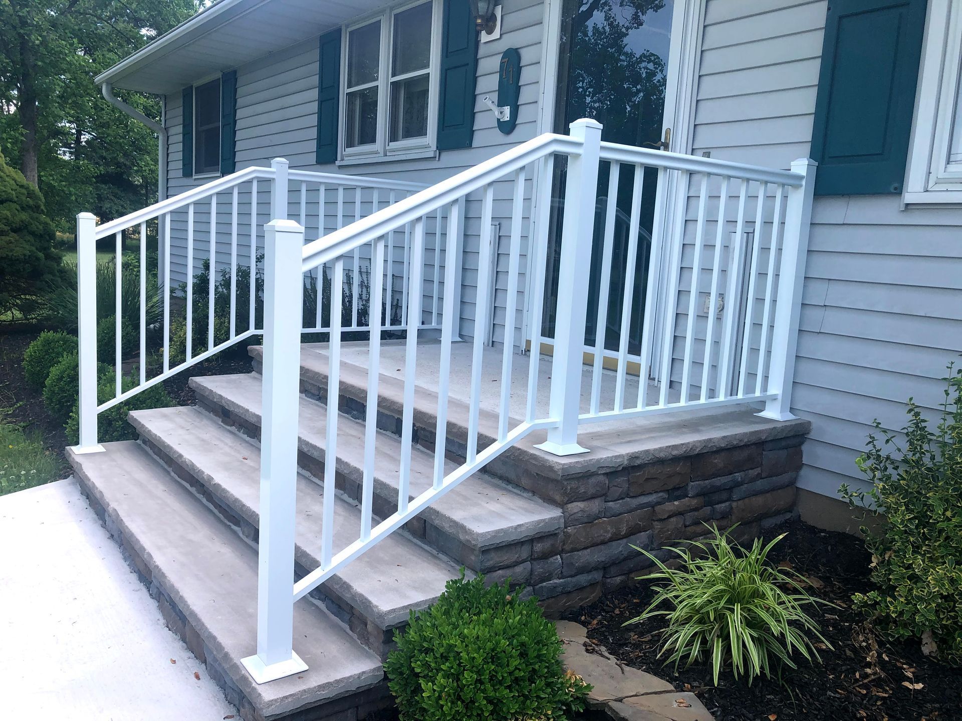 Front porch steps with white railing on a gray house, leading to an entryway with small shrubs below.