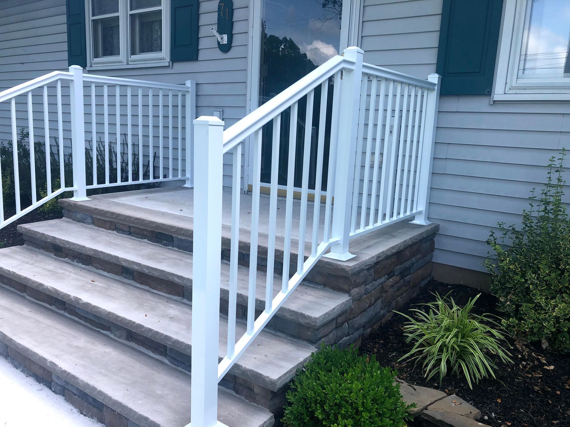 Front steps with white railings leading to a gray house entryway, with a small shrub beside the porch