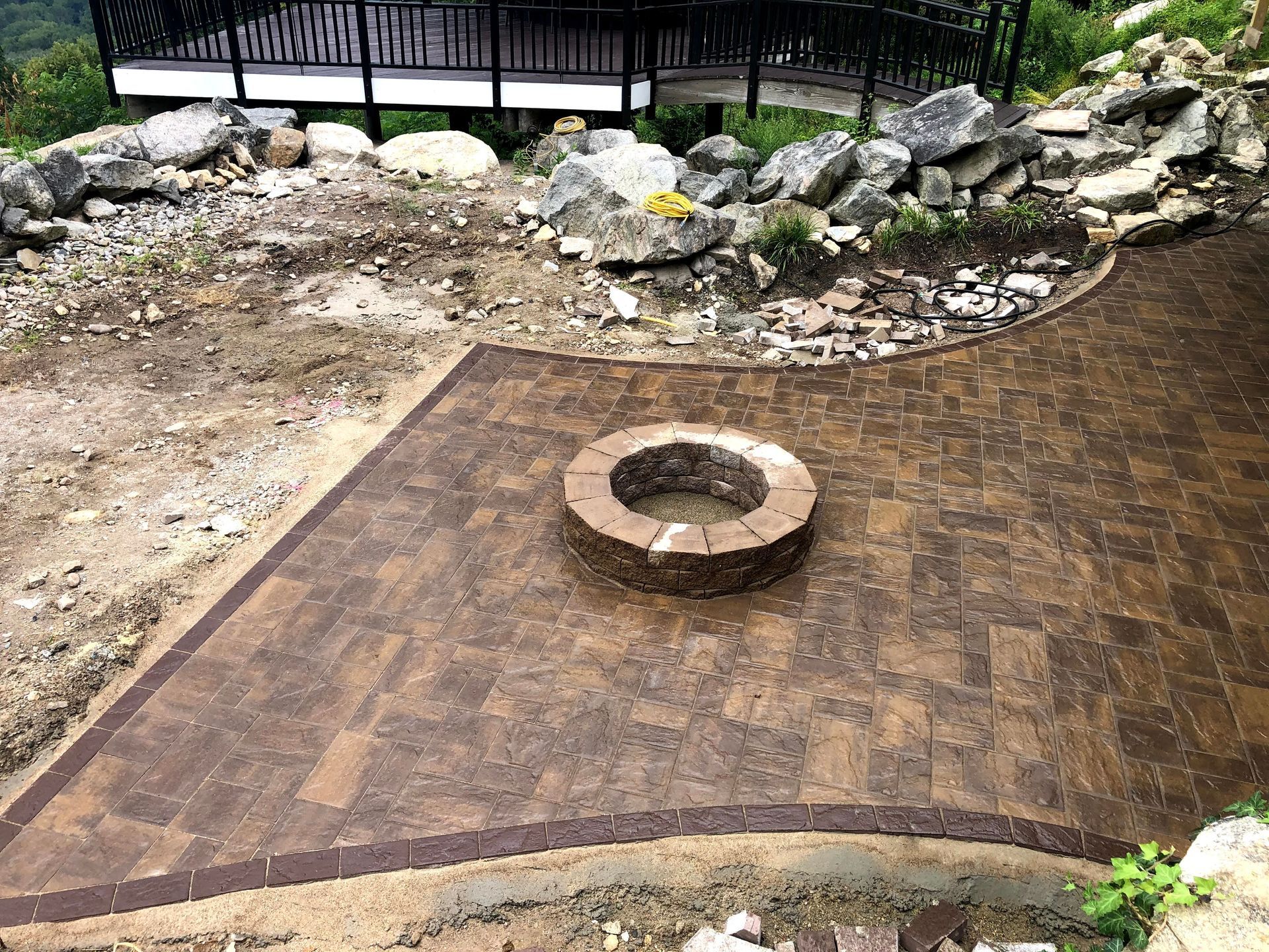 Stone fire pit on a brick patio beside a rocky landscaped area and black railing.