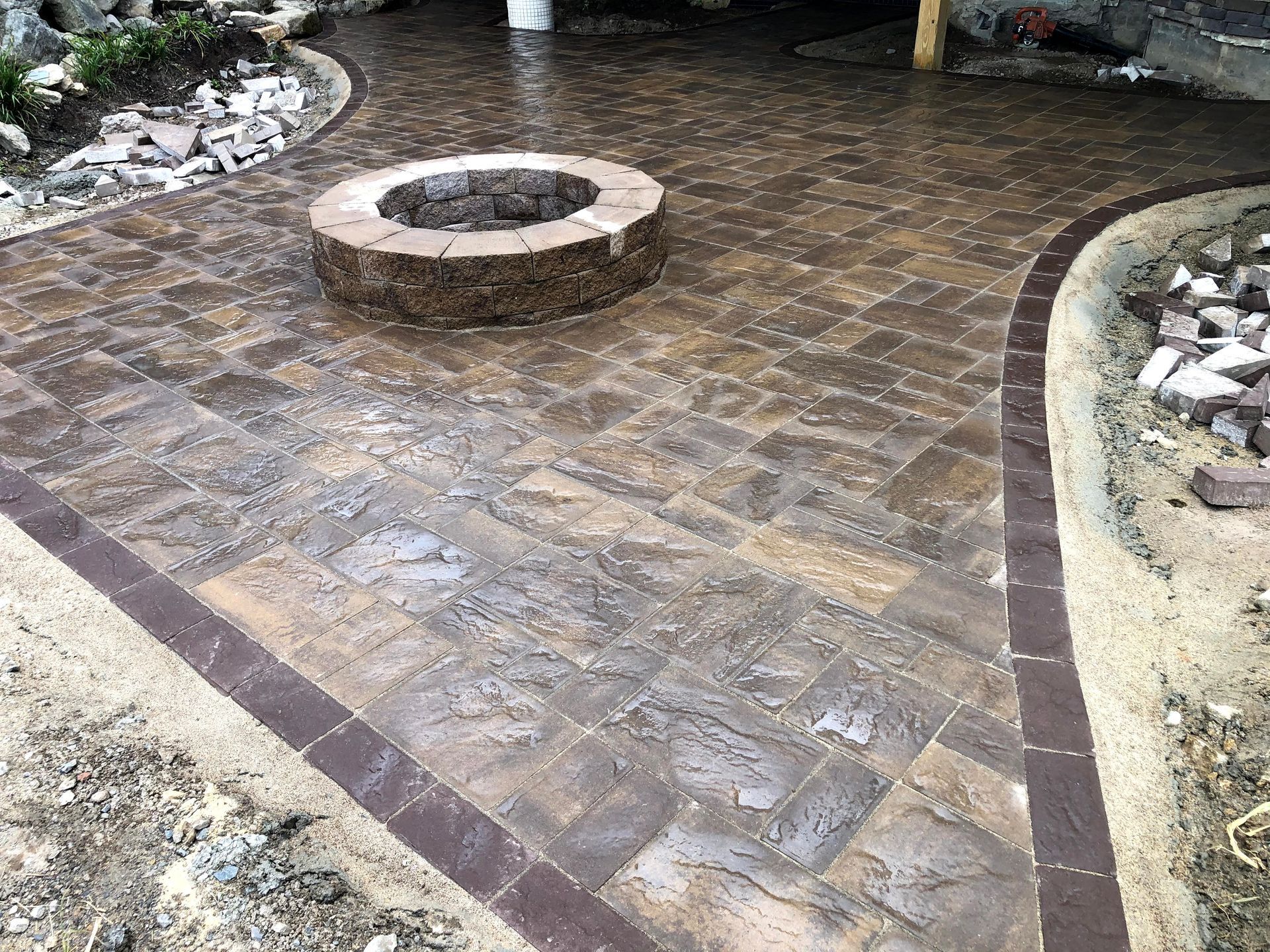 Brick-paved patio with a circular fire pit and curved dark border beside rocks and landscaping.