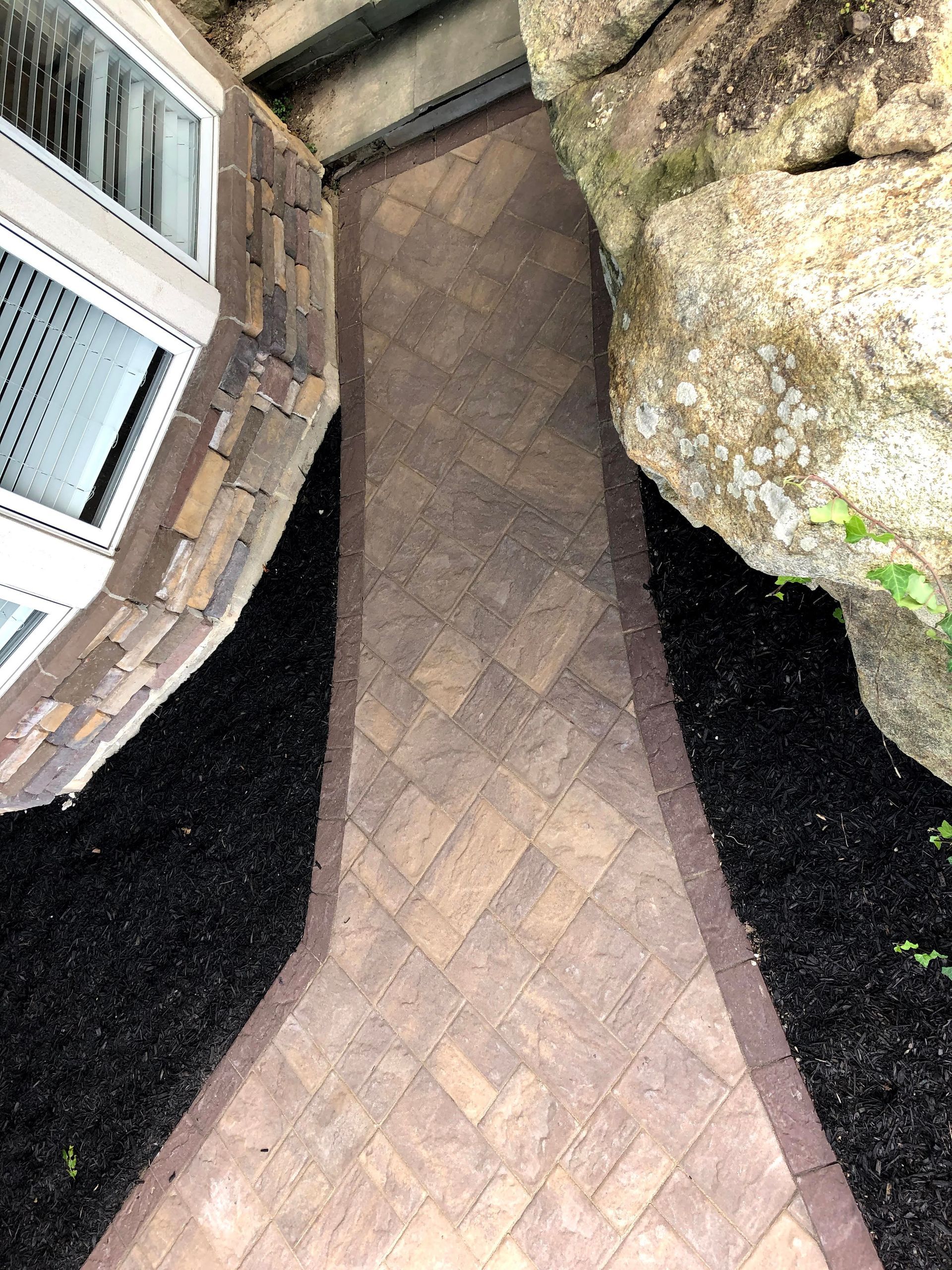 Narrow brick walkway between a house wall and large rocks, bordered by black gravel mulch.
