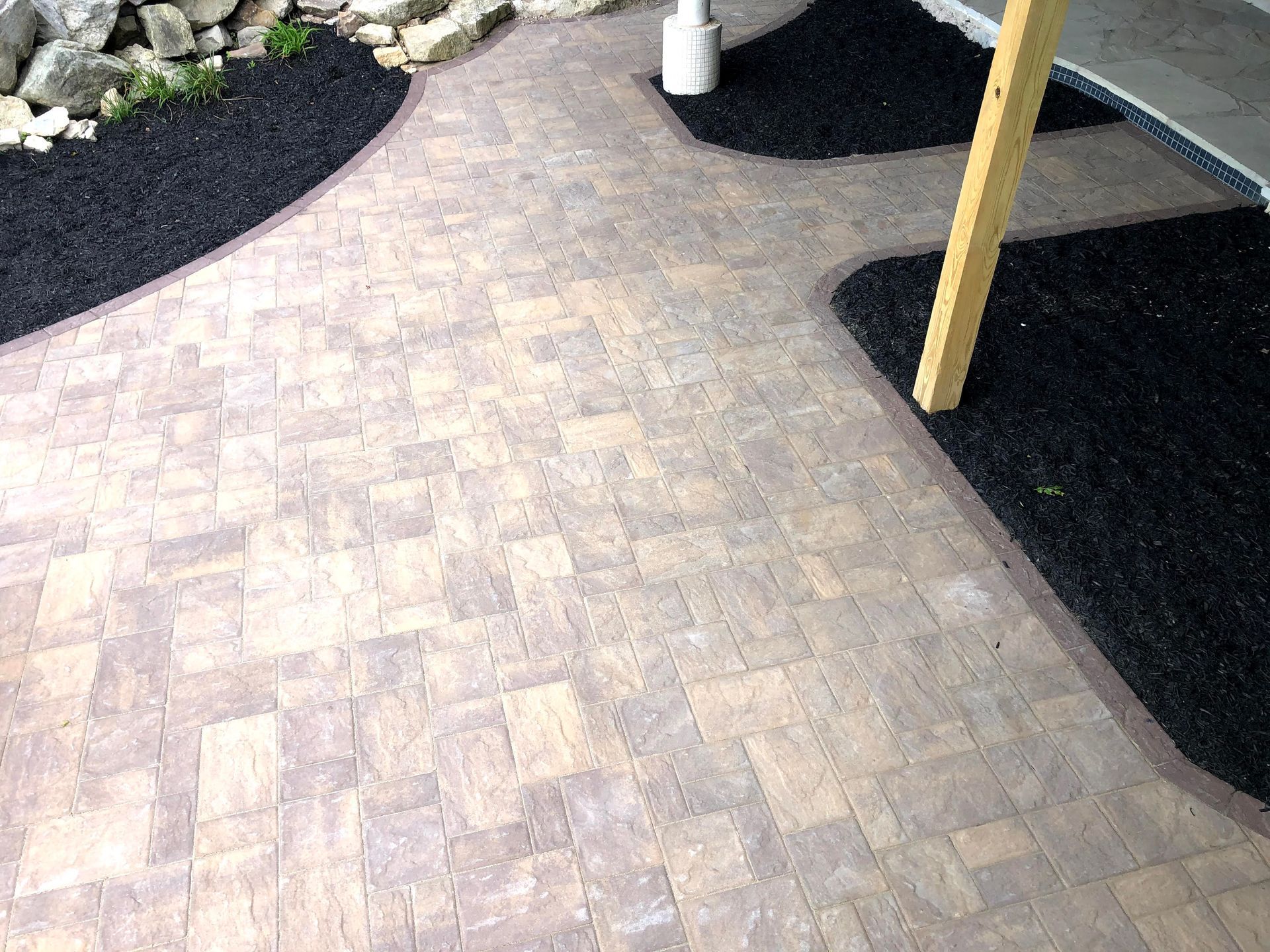 Brick paver walkway between black gravel beds with a wooden post and rocks рядом