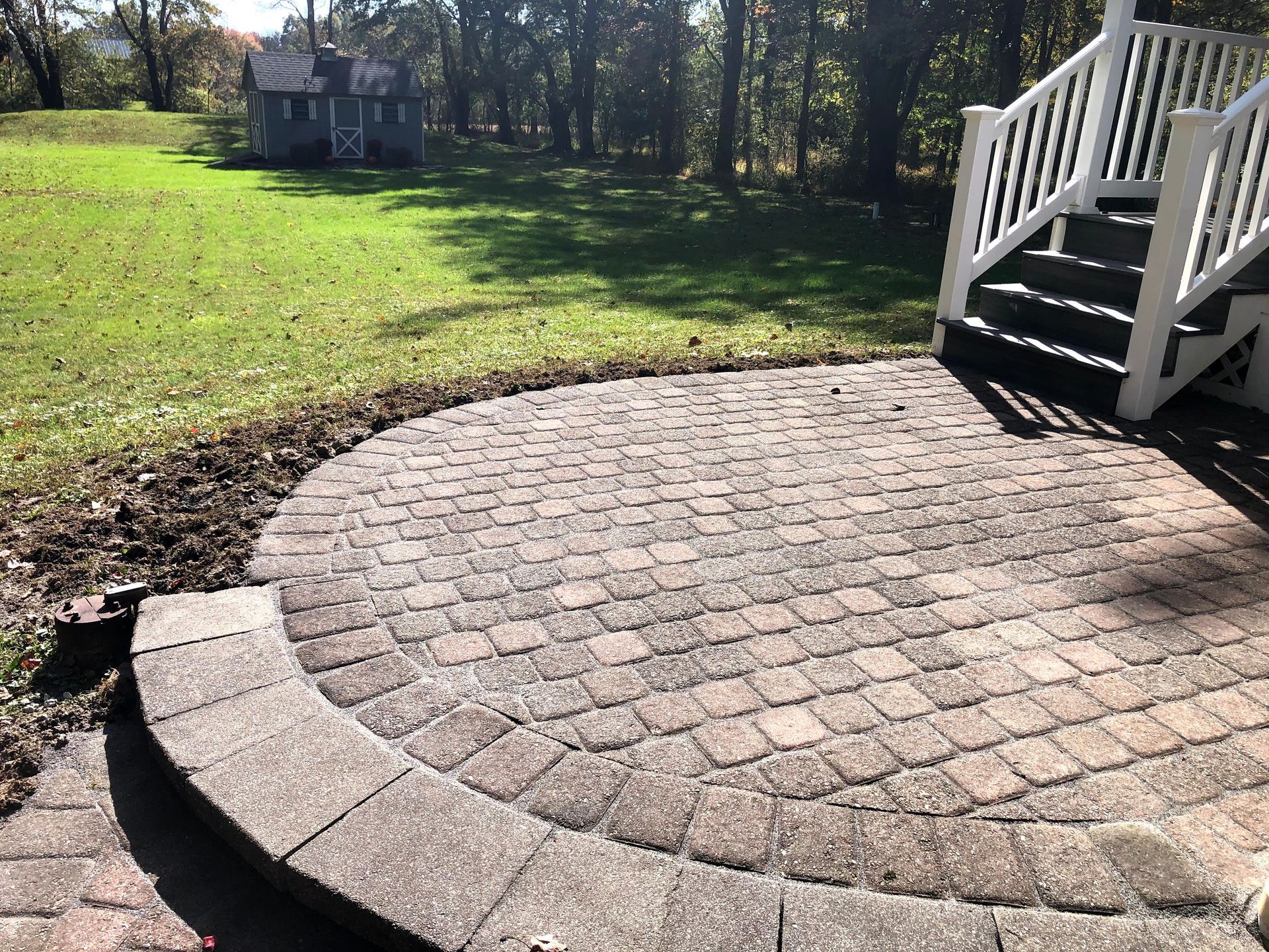 Curved brick patio beside white porch steps, with a grassy yard and trees in the background.