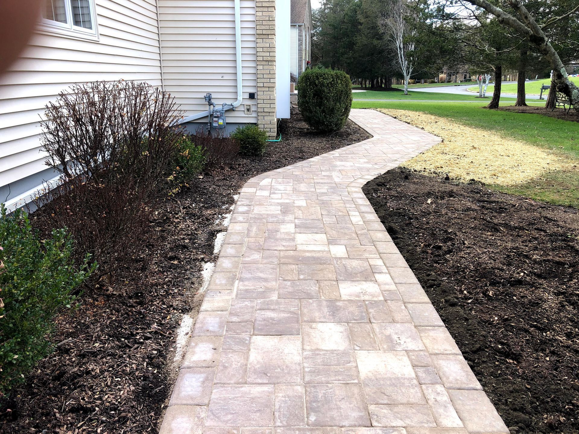 Curved stone walkway beside a house, bordered by shrubs and mulched garden beds.