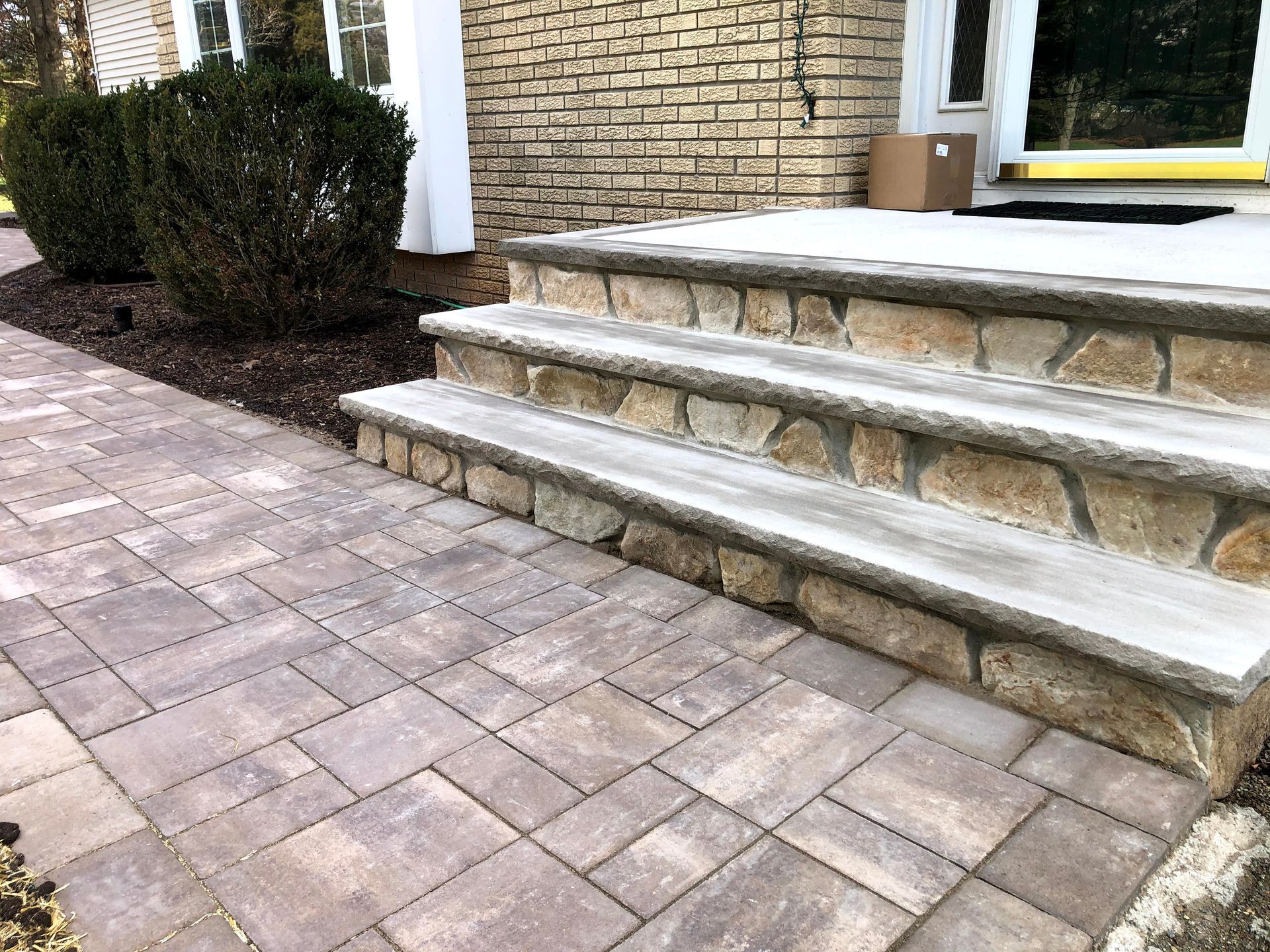Stone front steps beside a brick house and paved walkway, with a bush to the left.
