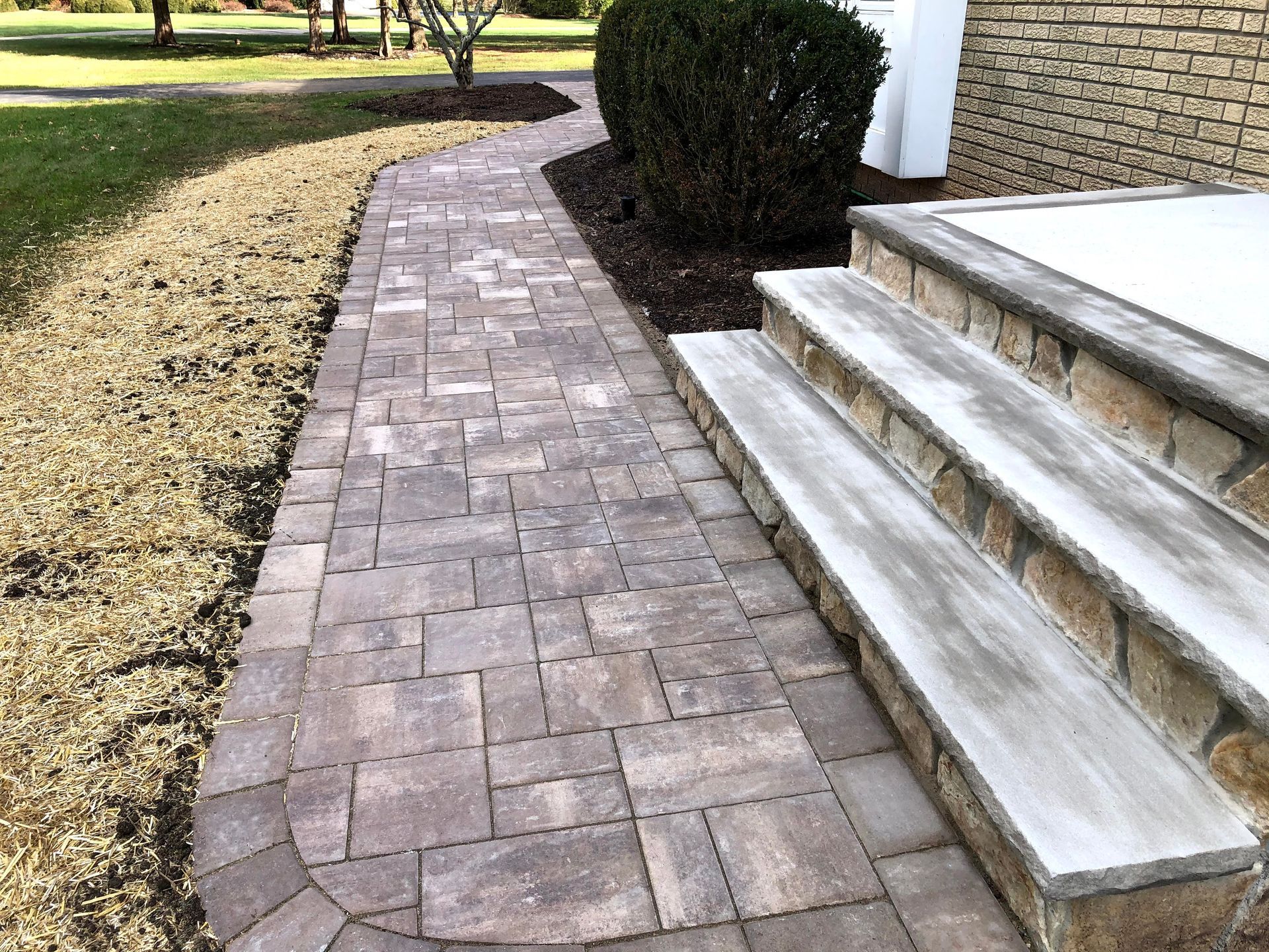 Curved brick walkway beside white stone steps and a trimmed hedge in a yard