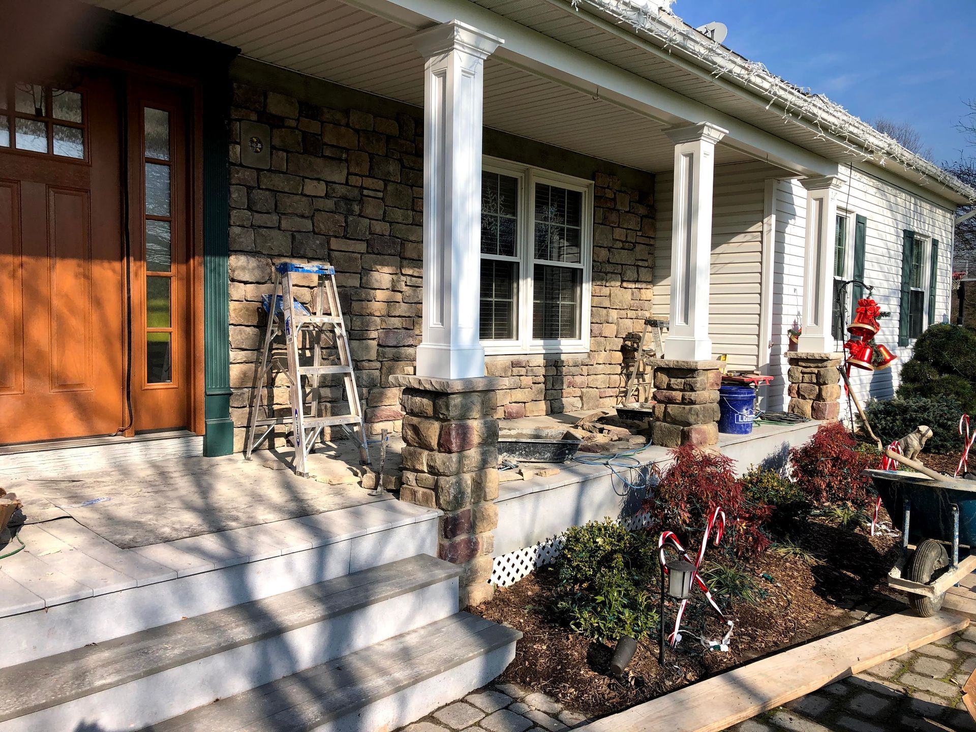 Front porch of a stone house with steps, white columns, wooden door, and flower beds along the walkway