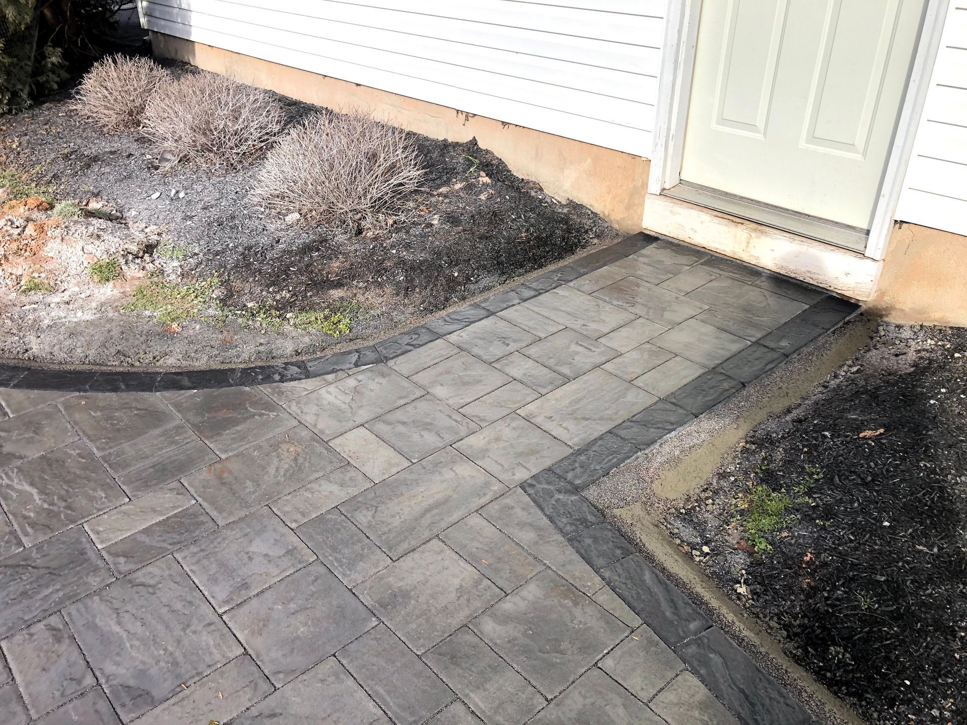 Paved walkway with gray pavers leading to a white front door beside mulch beds and a house wall