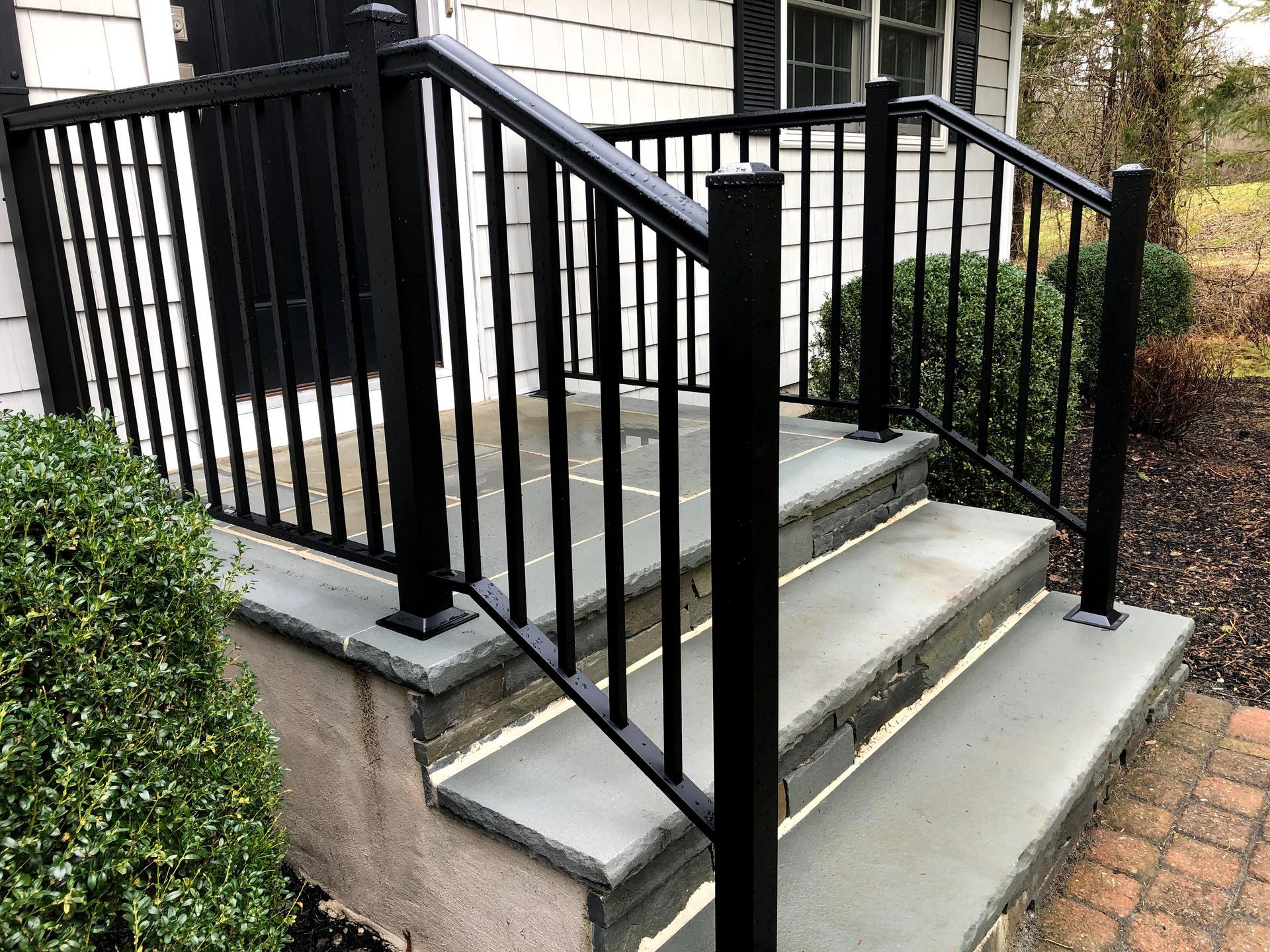 Black metal handrail and railings on concrete front steps beside a brick house.