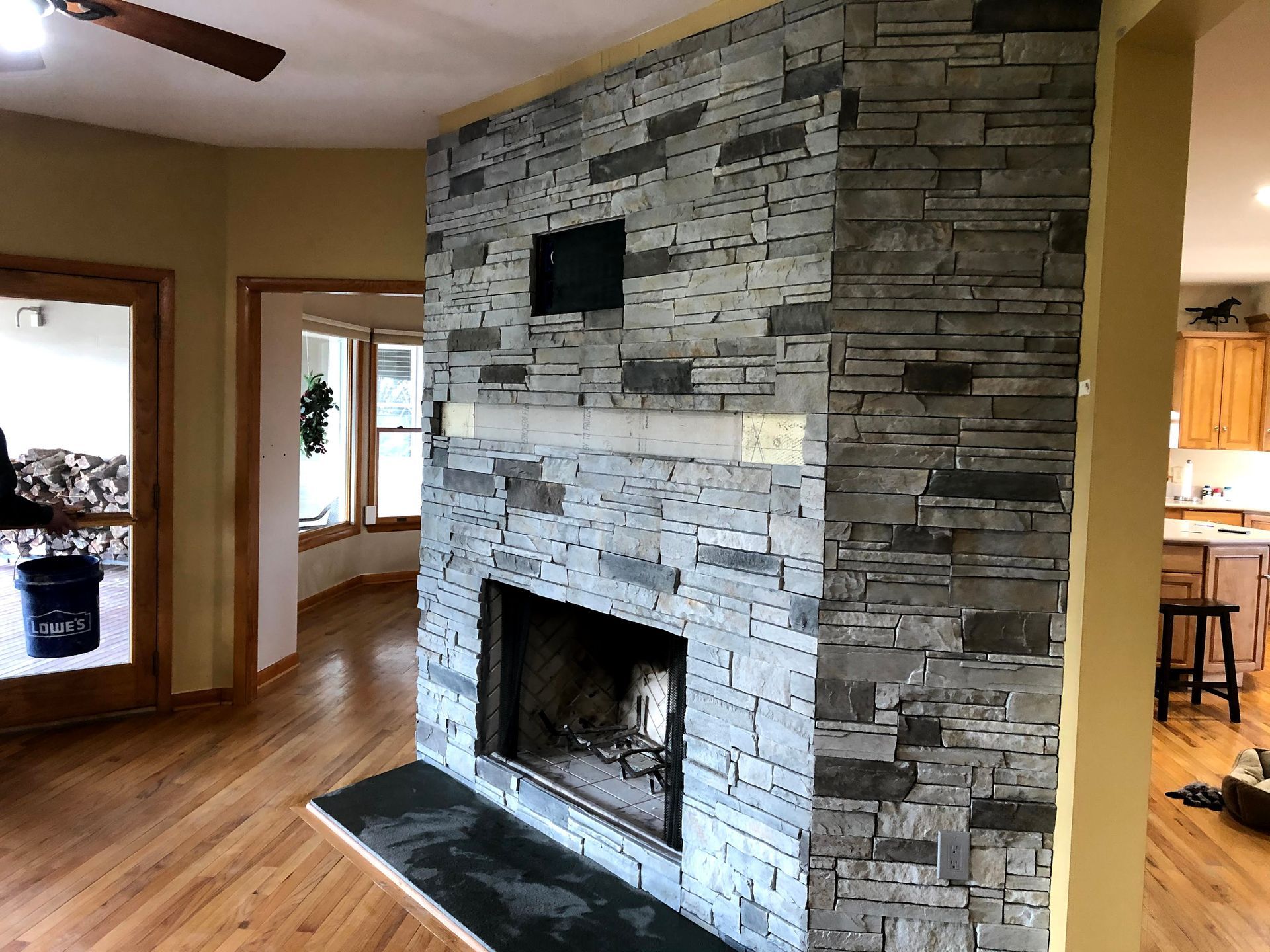 Living room with a stone fireplace wall and hardwood floors, open to a kitchen and dining area