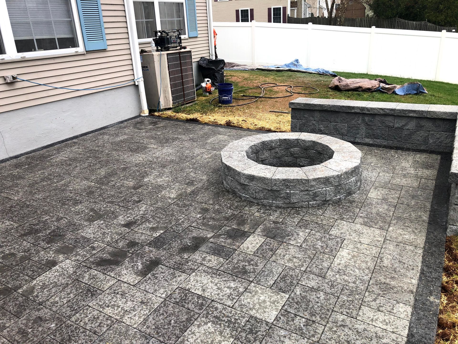 Backyard patio with a circular fire pit and stone pavers beside a house and white fence