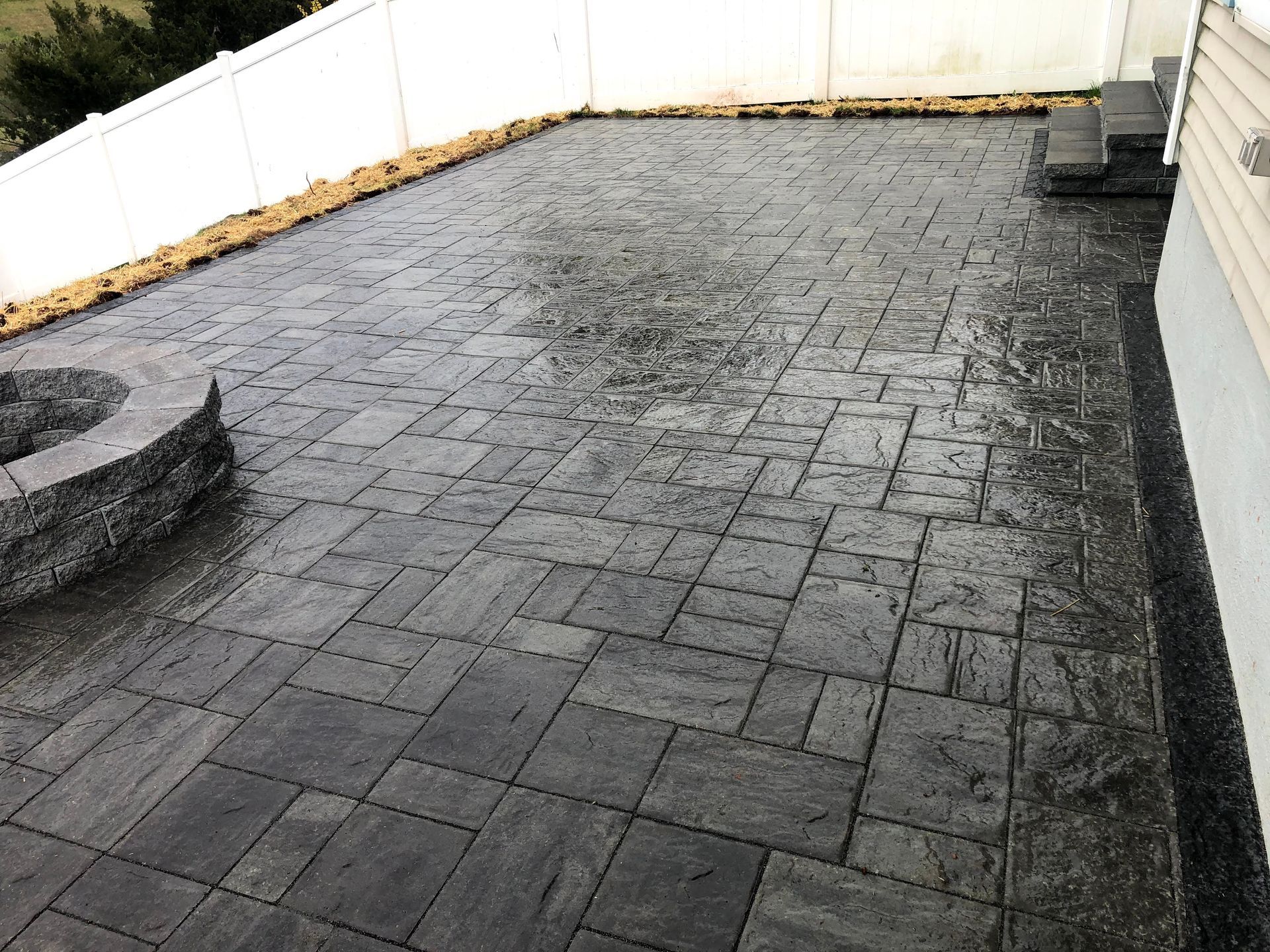 Empty paved patio with dark stone pavers and a white fence along the back and left sides.