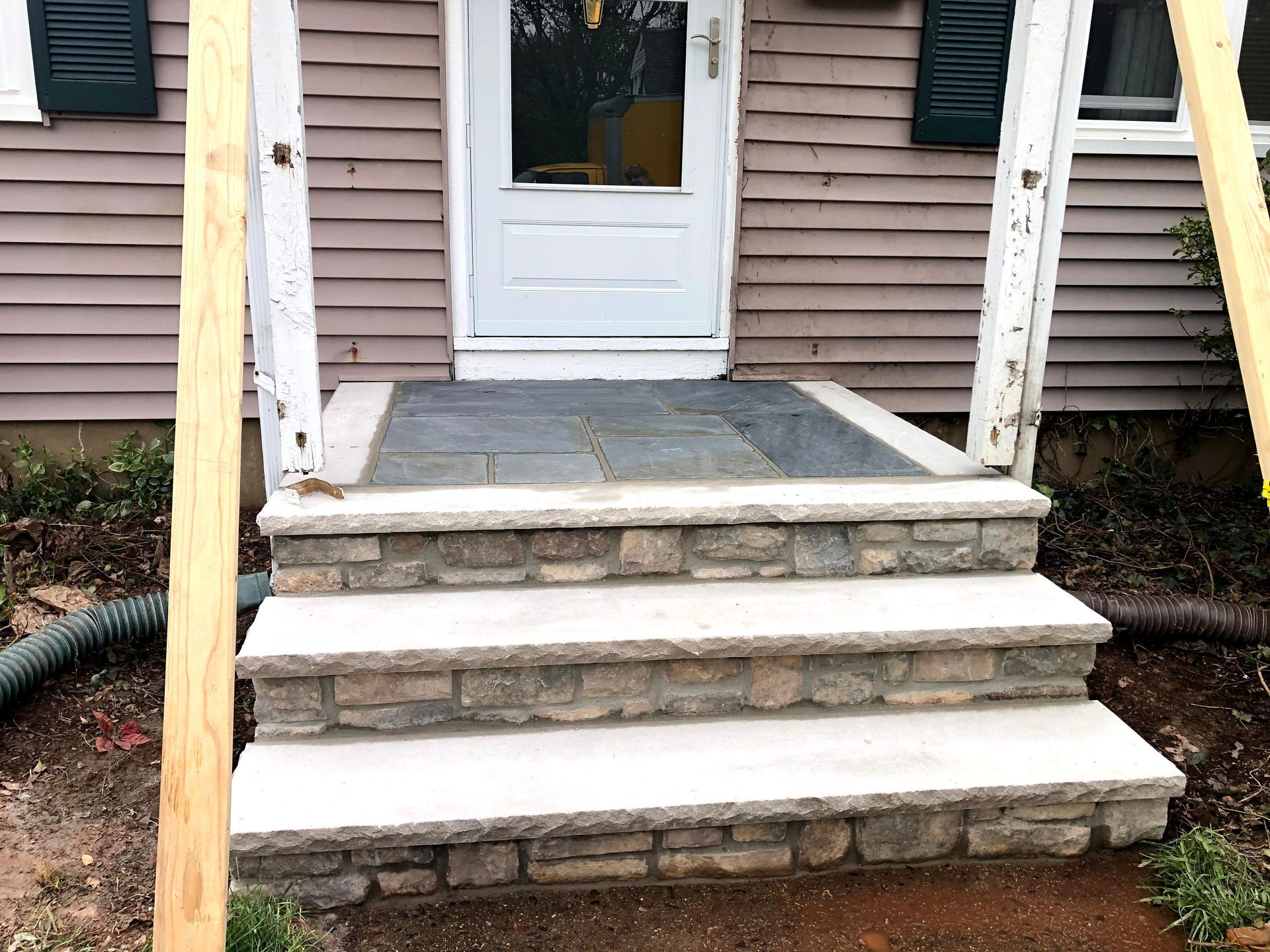 Front porch with four stone steps, white door, and wooden posts on a house exterior.