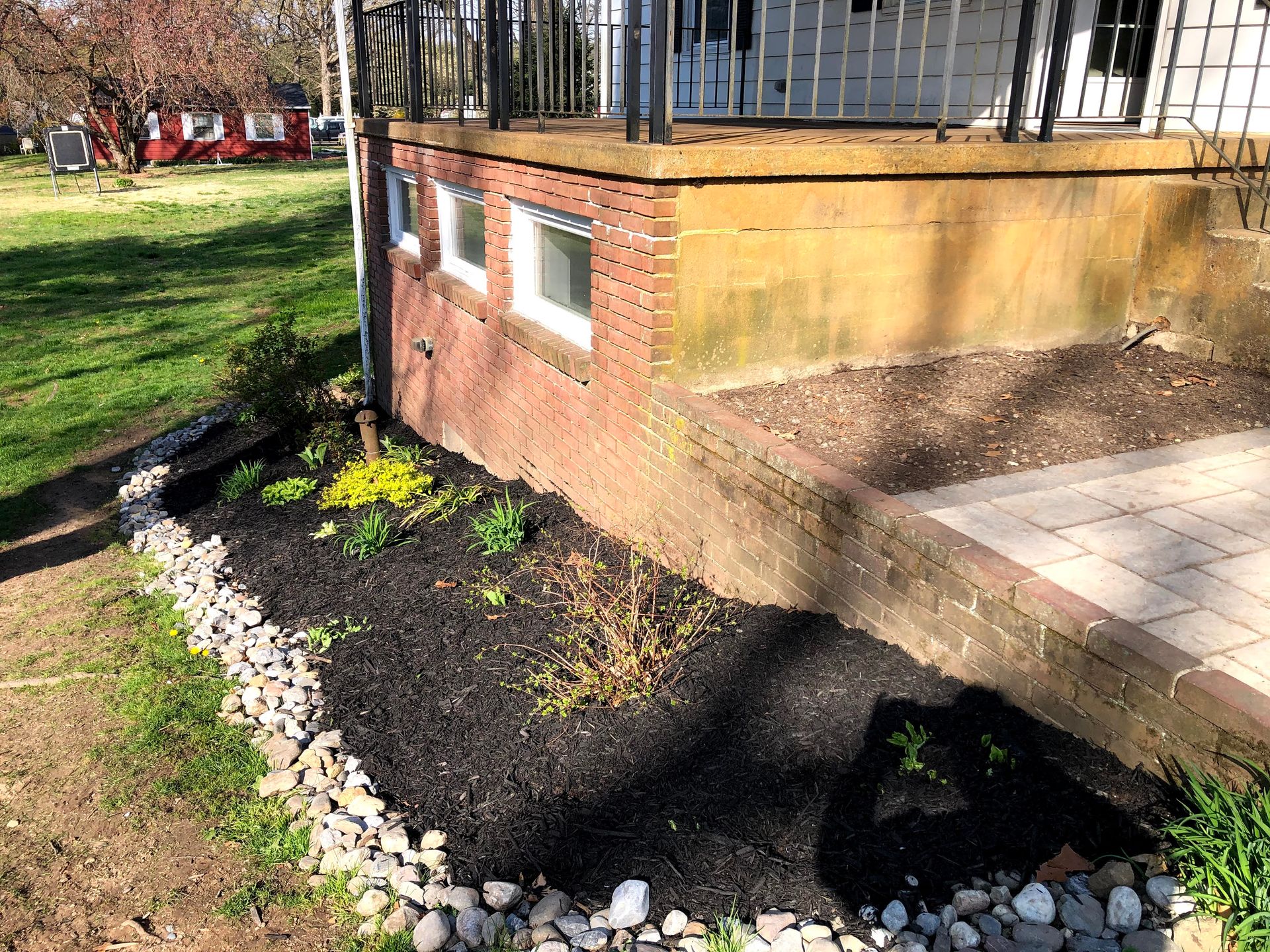 Brick house corner with white-trimmed windows, tiled patio, and a flower bed edged with white stones.