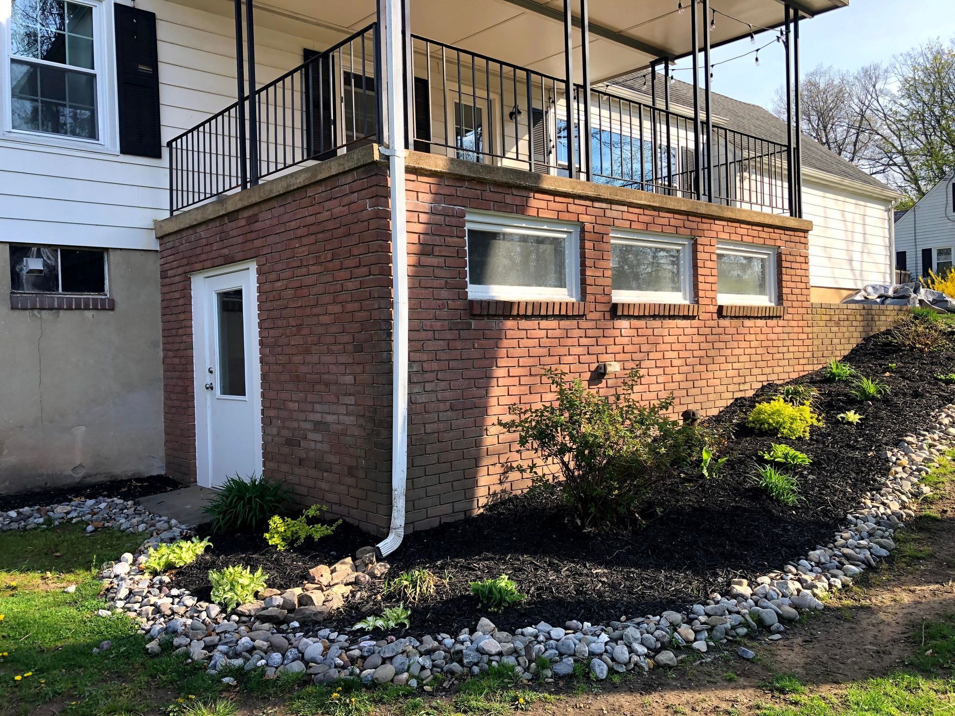 Brick home addition with white door and windows, black railing balcony, and landscaped rock border