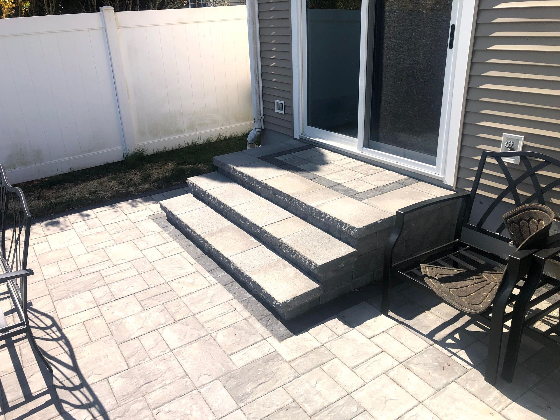 Back patio with three concrete steps leading to a glass door, bordered by a white fence and outdoor furniture.