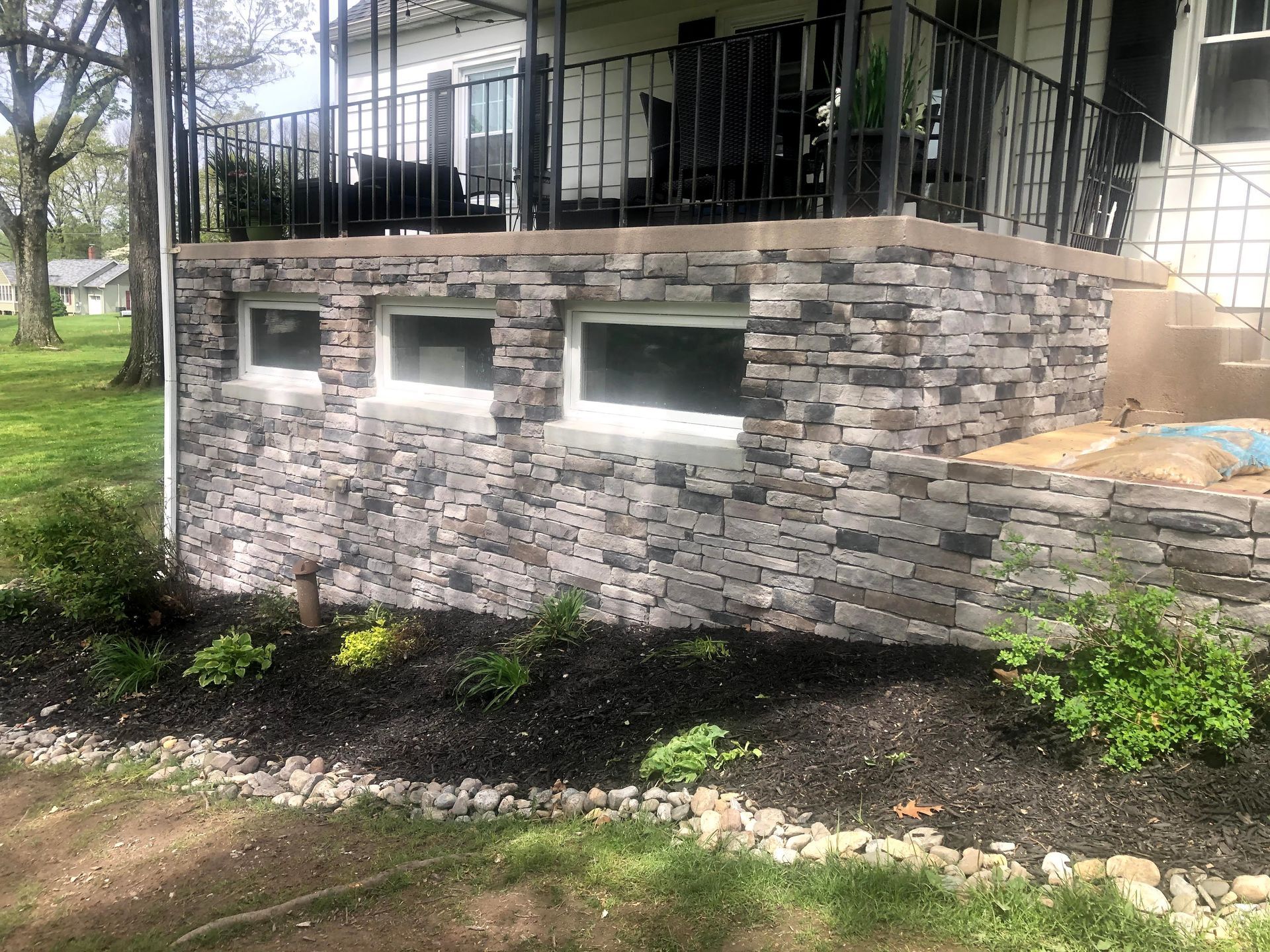 Stone retaining wall with three windows beside a house, under a black metal patio railing.