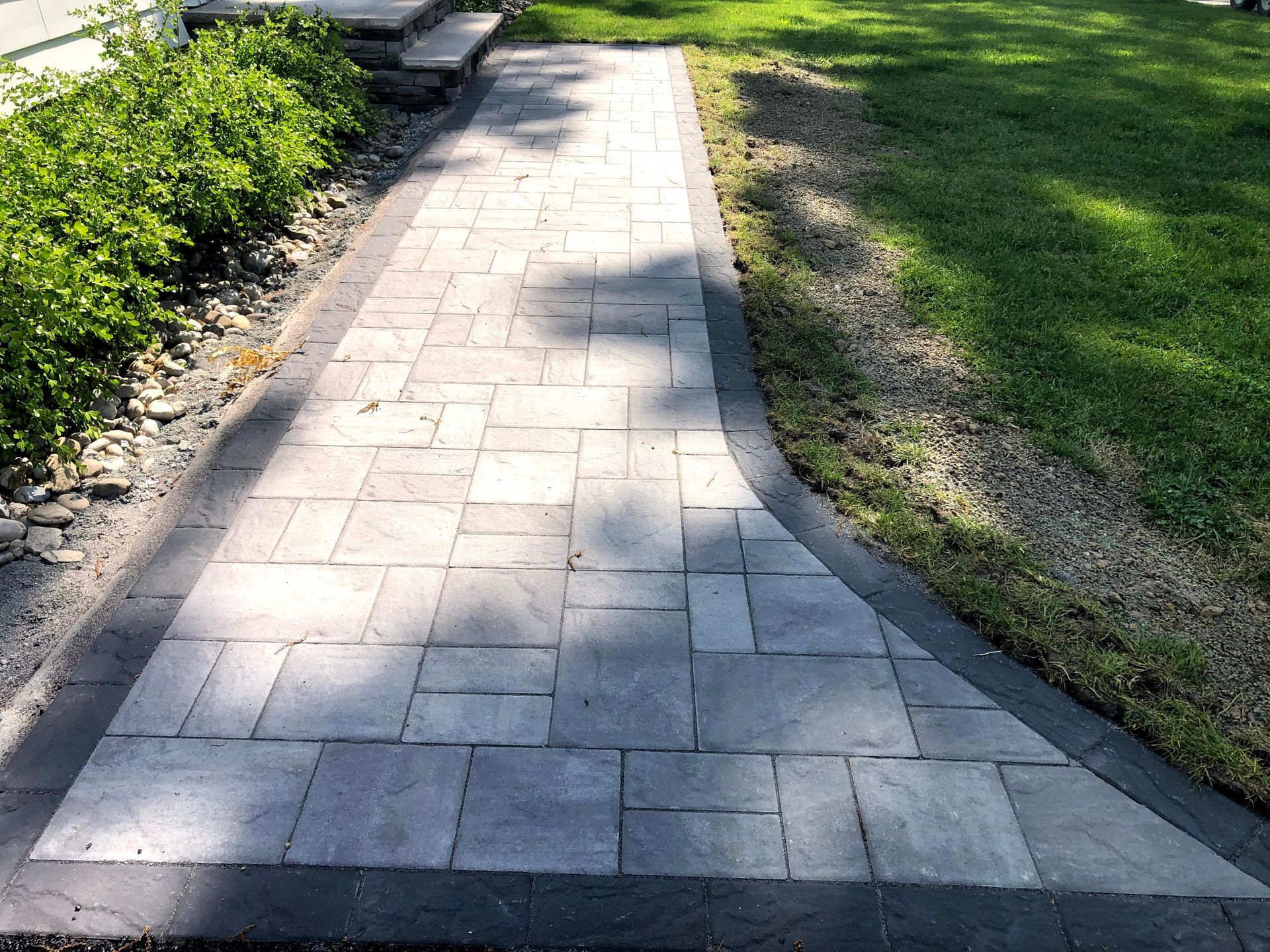 Sunlit stone walkway curving through a grassy park, with shrubs and benches along the side