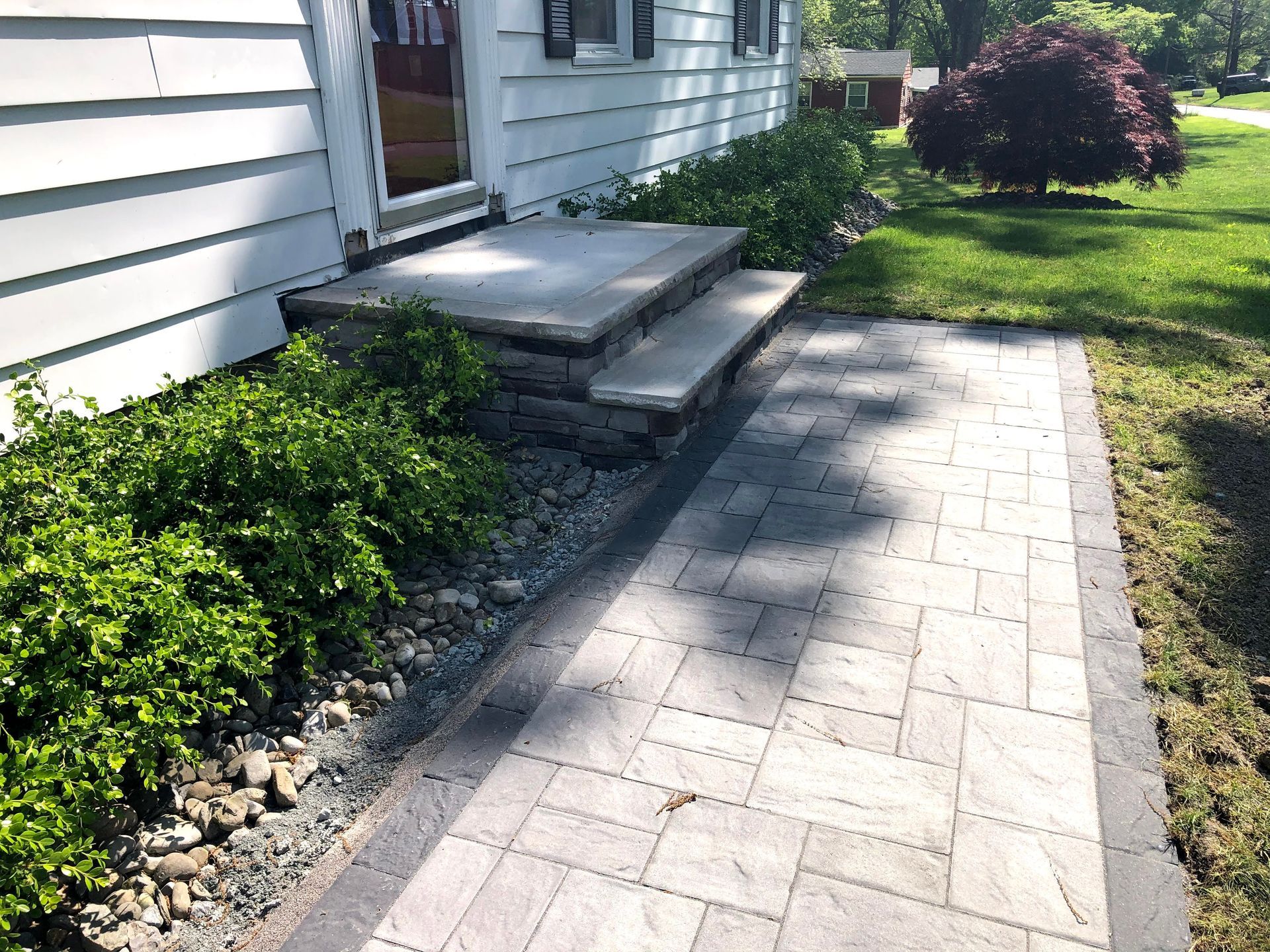 Brick walkway beside a white house, leading to a small front porch with steps and a green lawn.