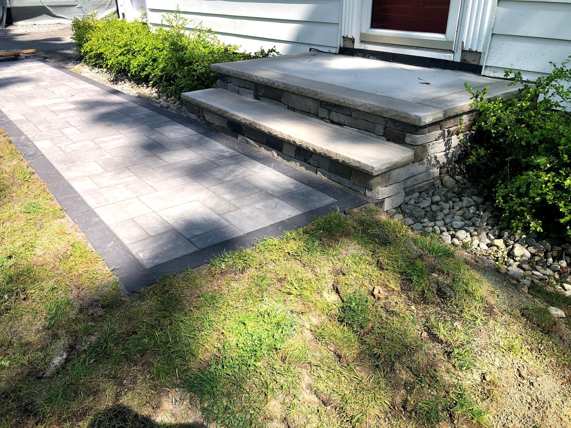 Front steps and concrete walkway beside a white house, bordered by grass and shrubs