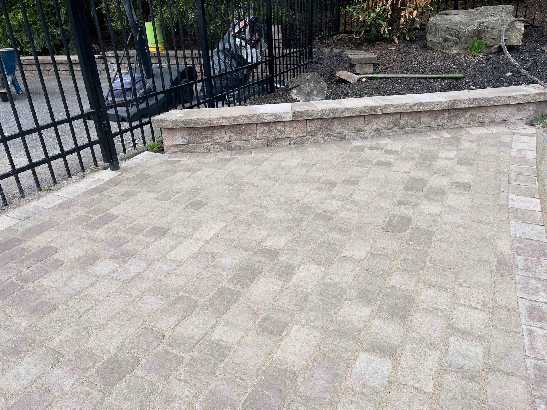 Paved courtyard beside a black metal fence and stone steps, with a low retaining wall in the background
