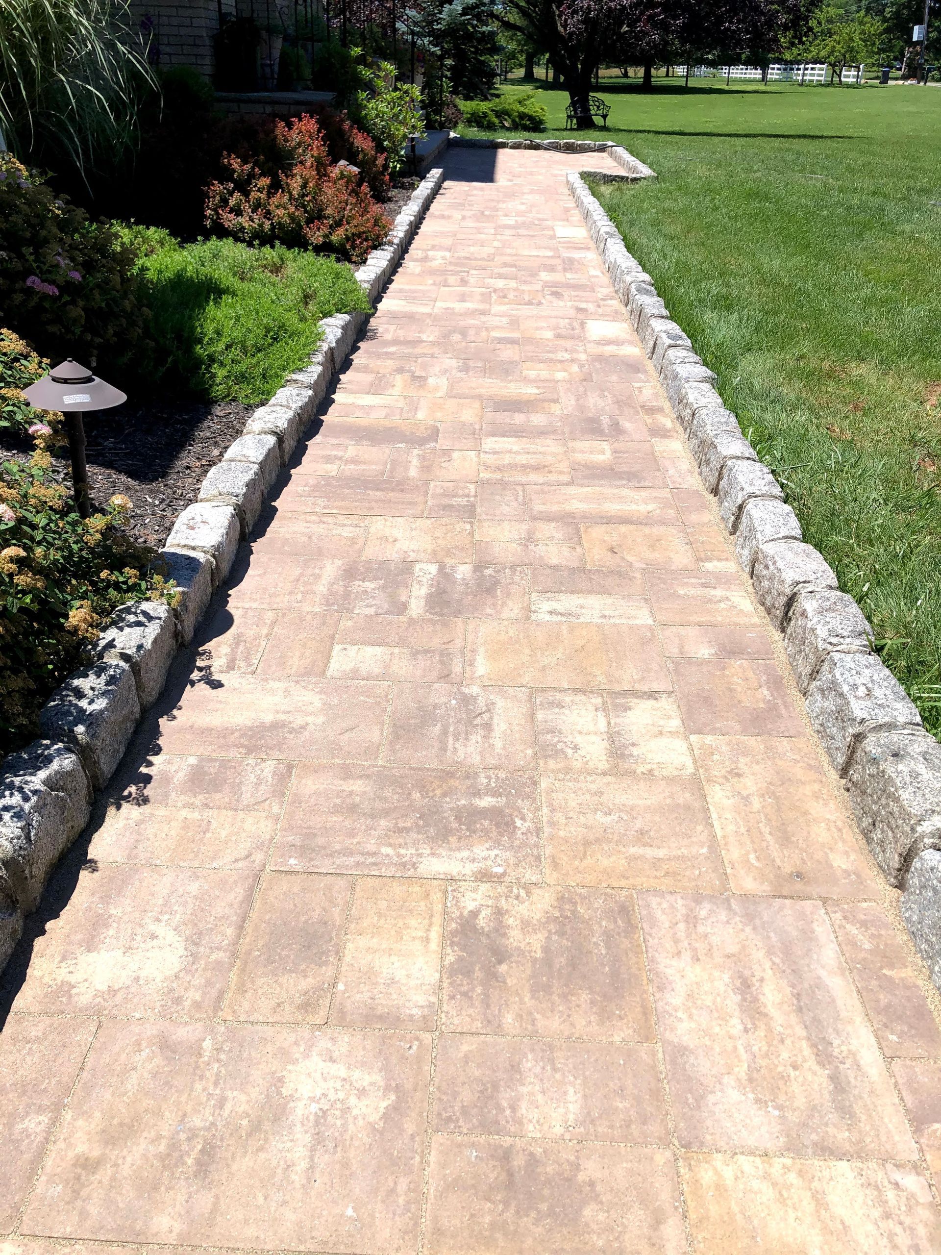 Stone walkway through a green garden, bordered by rocks and shrubs, leading into the distance.