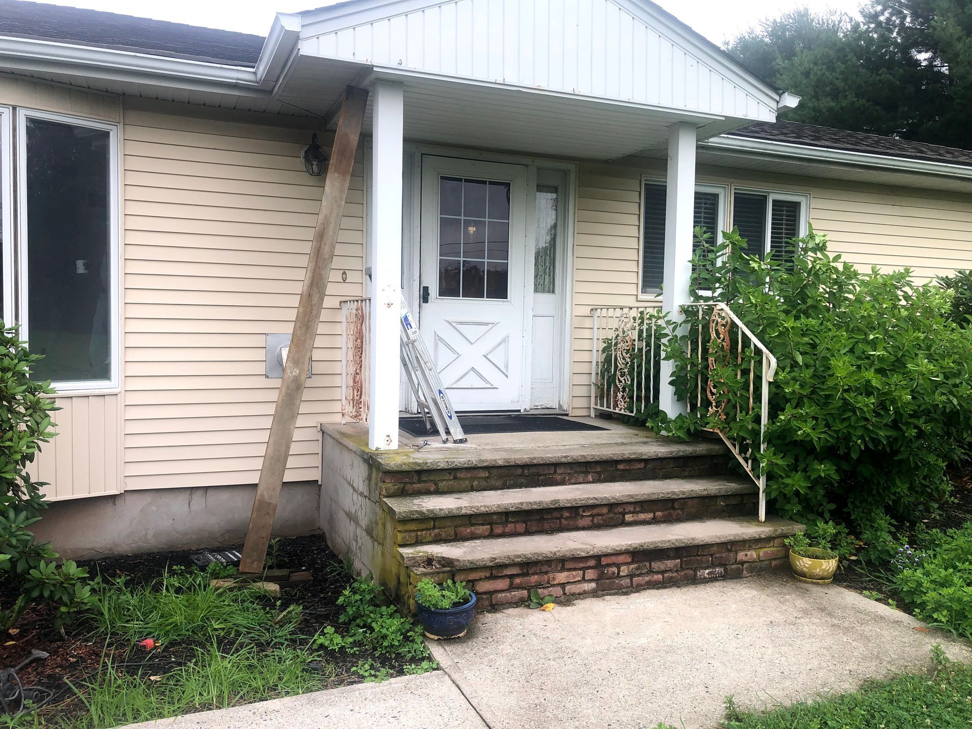 Front entrance of a beige house with steps, porch, white door, and overgrown bushes on the right