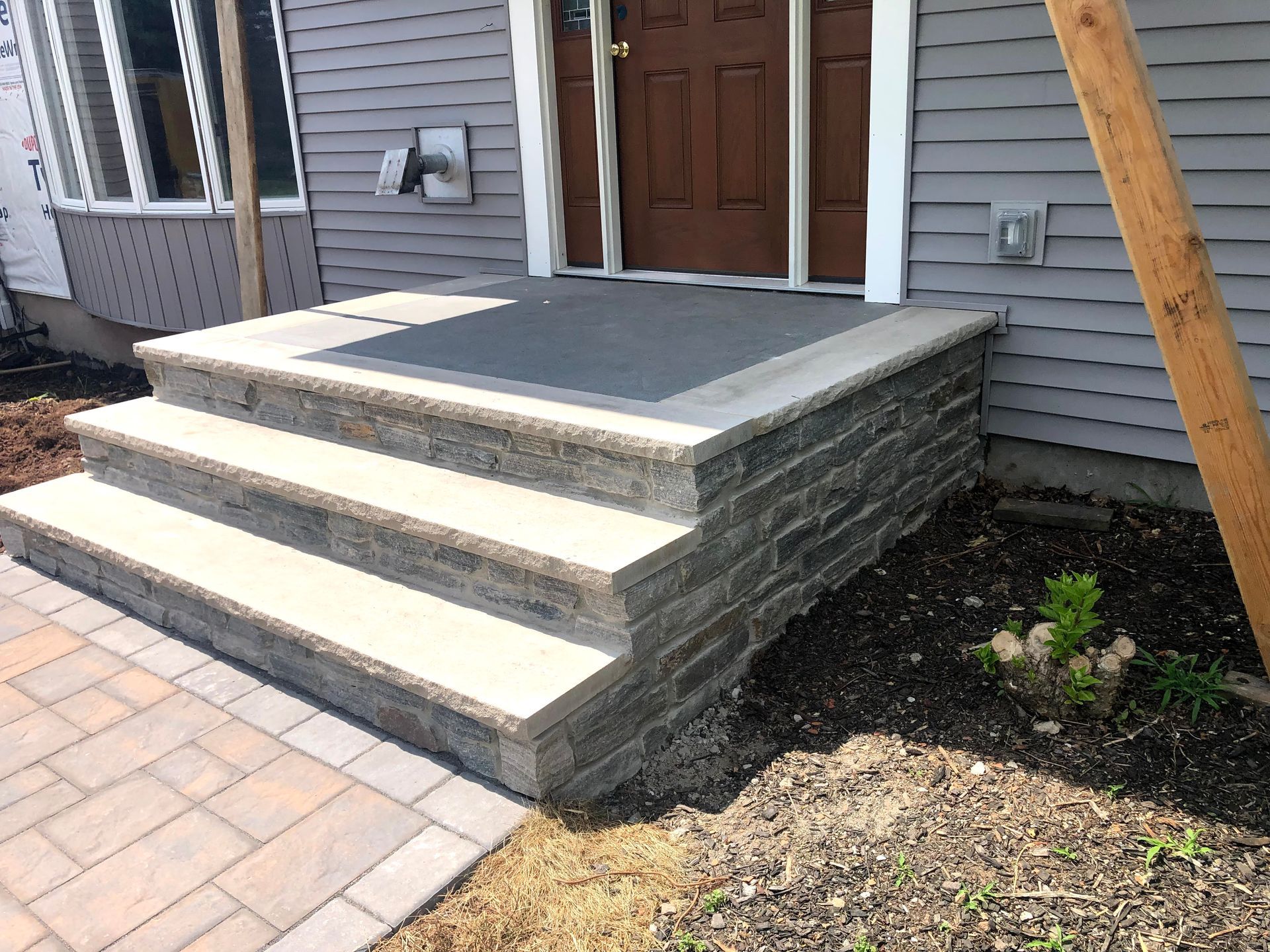 Front porch with two concrete steps, stone sidewalls, and a wooden front door on a gray house