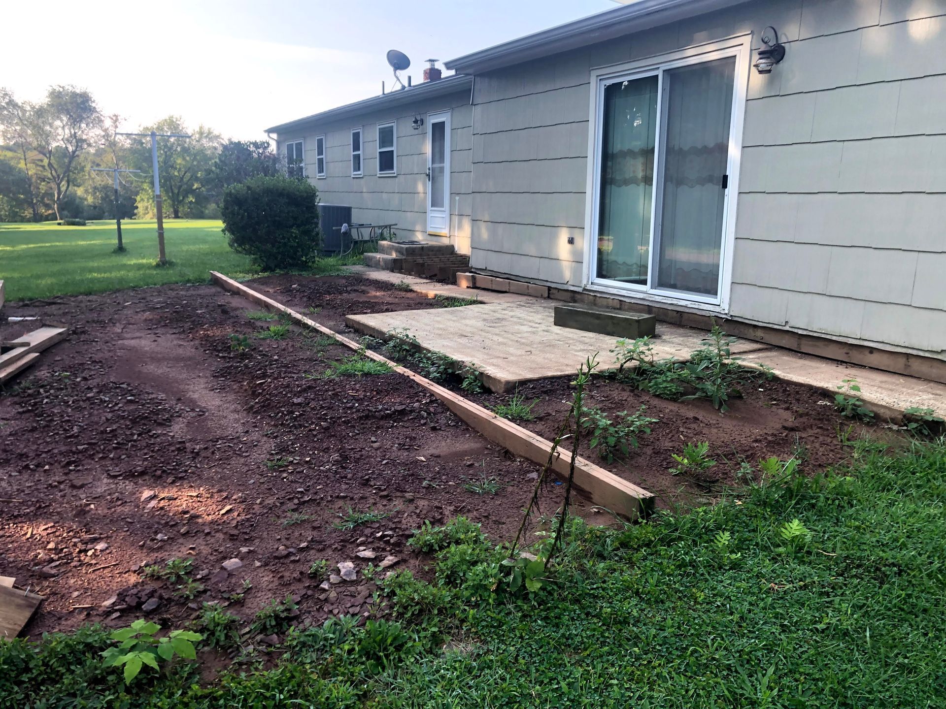 Backyard patio and garden bed beside a beige house, with sliding glass doors and a grassy lawn