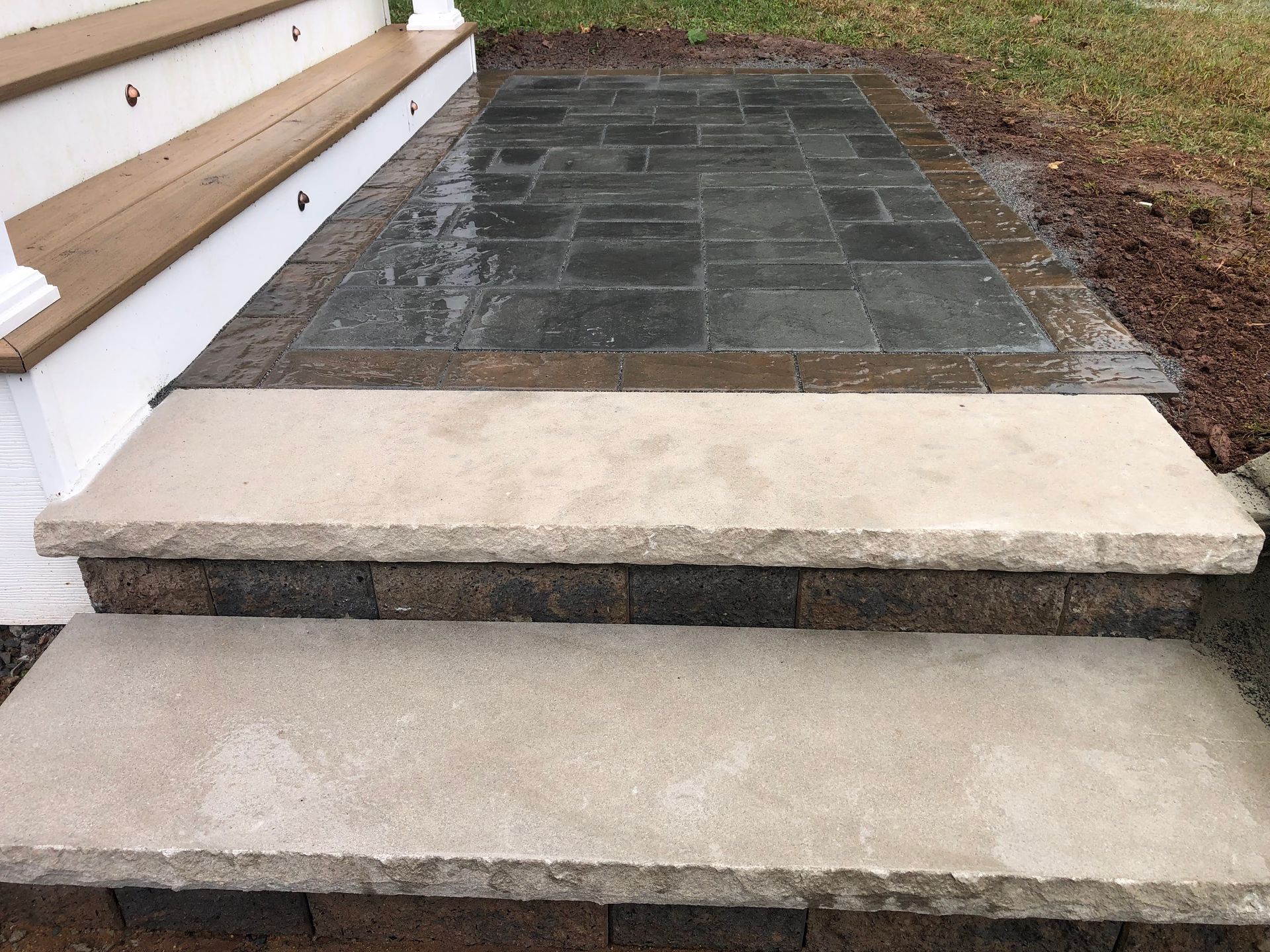 Stone steps leading to a raised patio with dark pavers and a white wall with wood accents