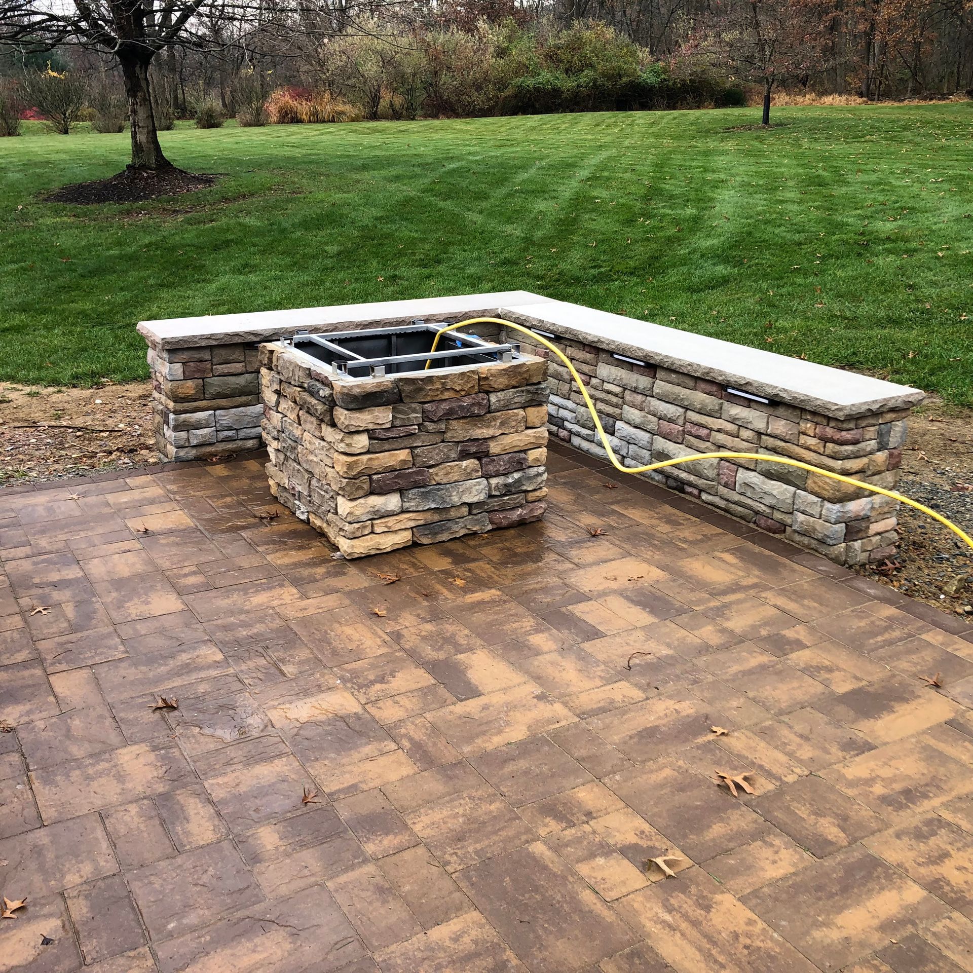Stone outdoor fire pit with built-in bench seating on a brick patio beside a grassy yard