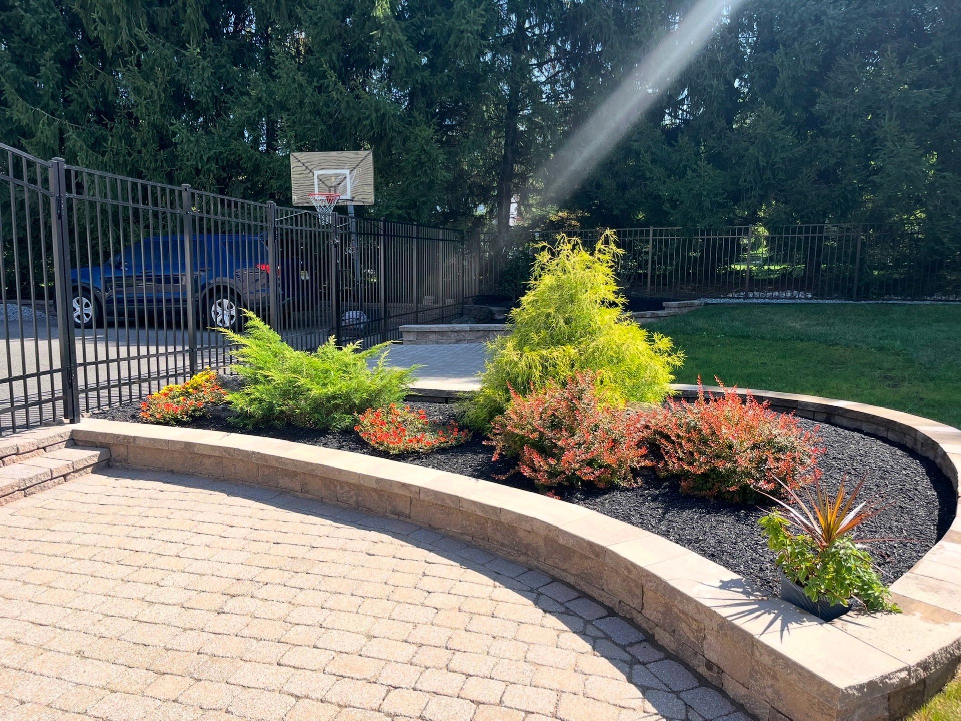 Curved brick planter with colorful shrubs beside a fenced basketball court in a sunny park