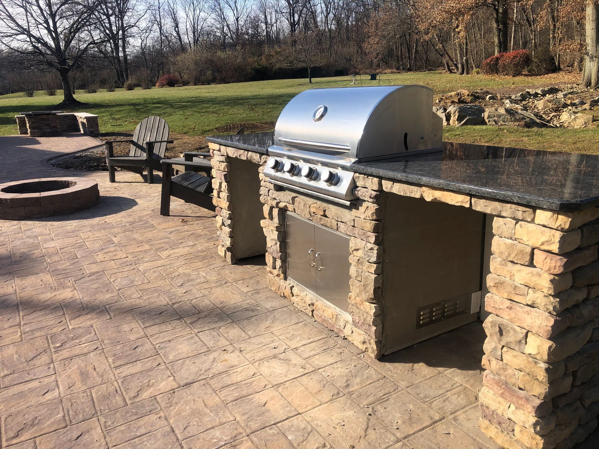 Outdoor stone grill and patio seating in a sunny backyard, with trees and a grassy lawn beyond.
