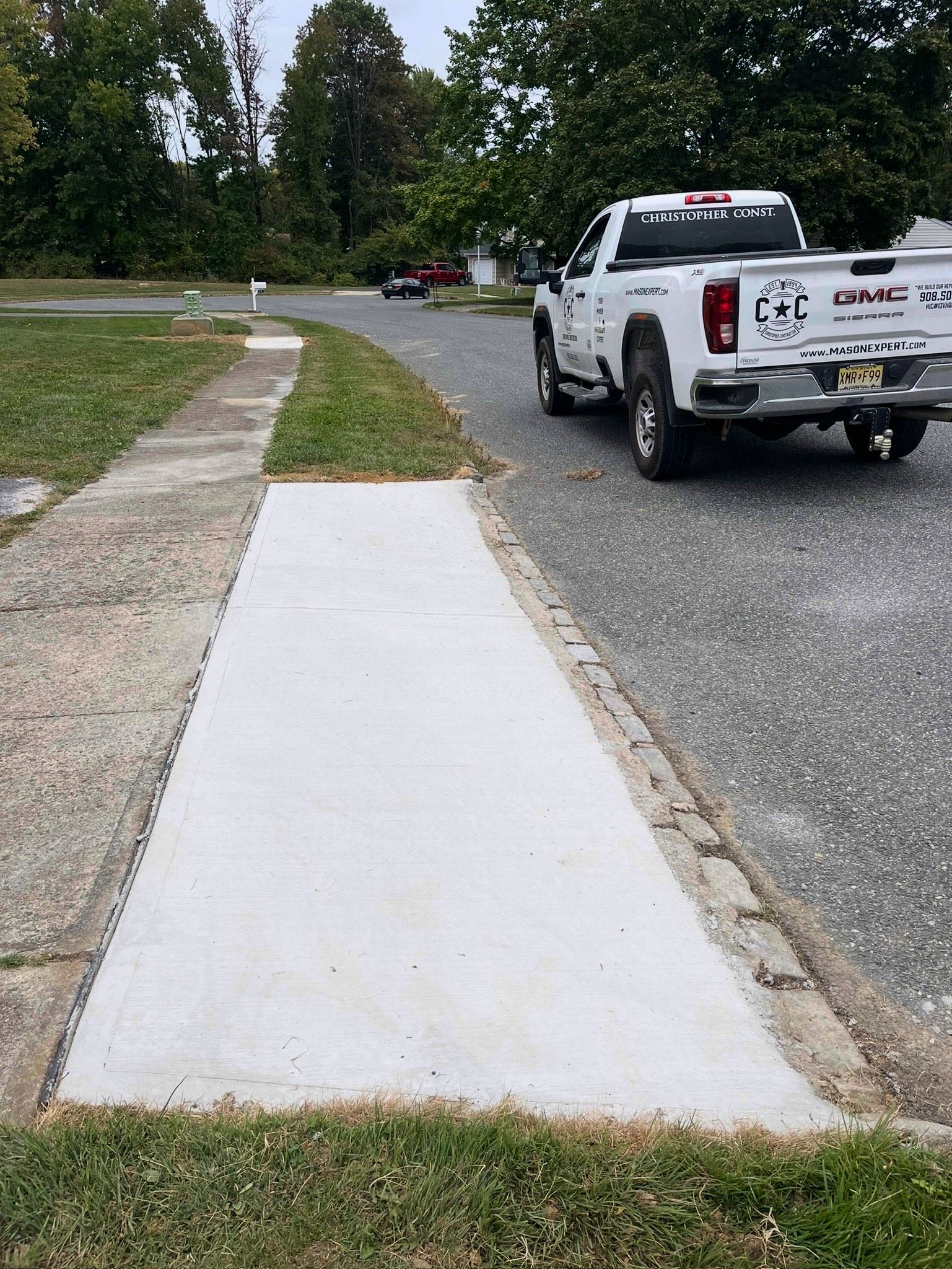 Fresh concrete sidewalk beside a gravel road, with a white pickup truck parked ahead under trees.