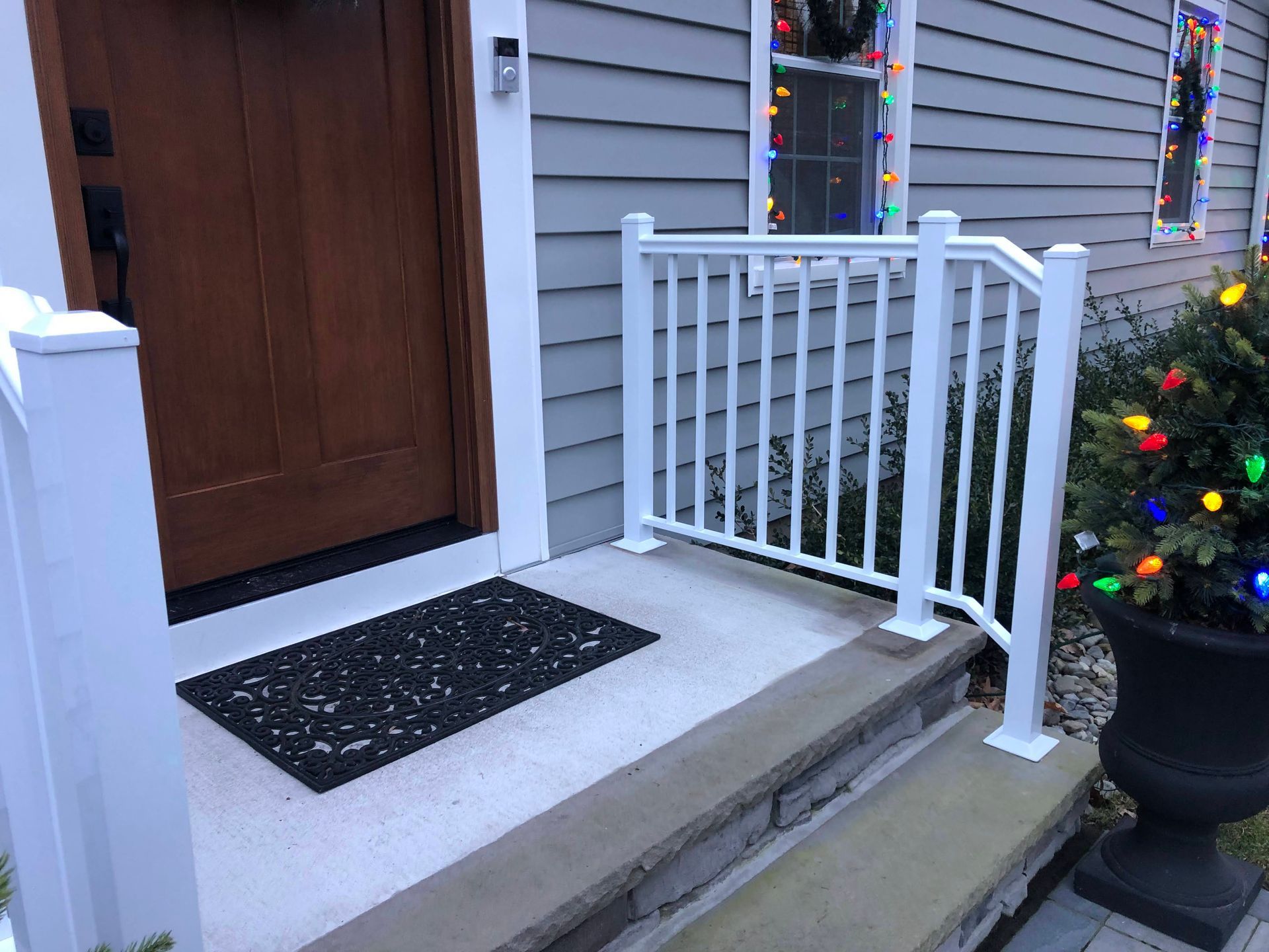 Front porch with a wooden door, white railing, concrete steps, and holiday lights by a large planter.