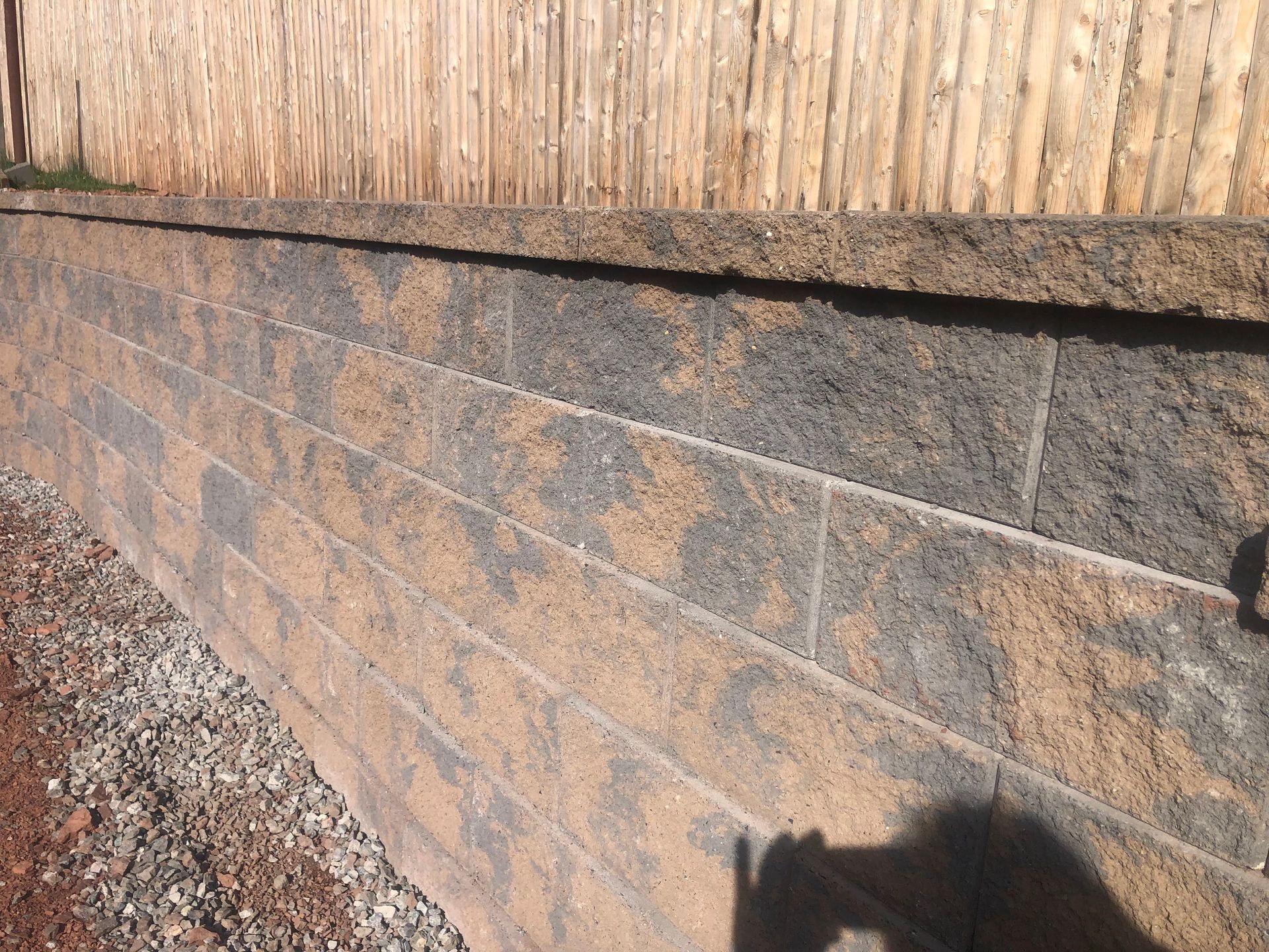 Stone retaining wall with a tan fence above and gravel along the base in sunlight