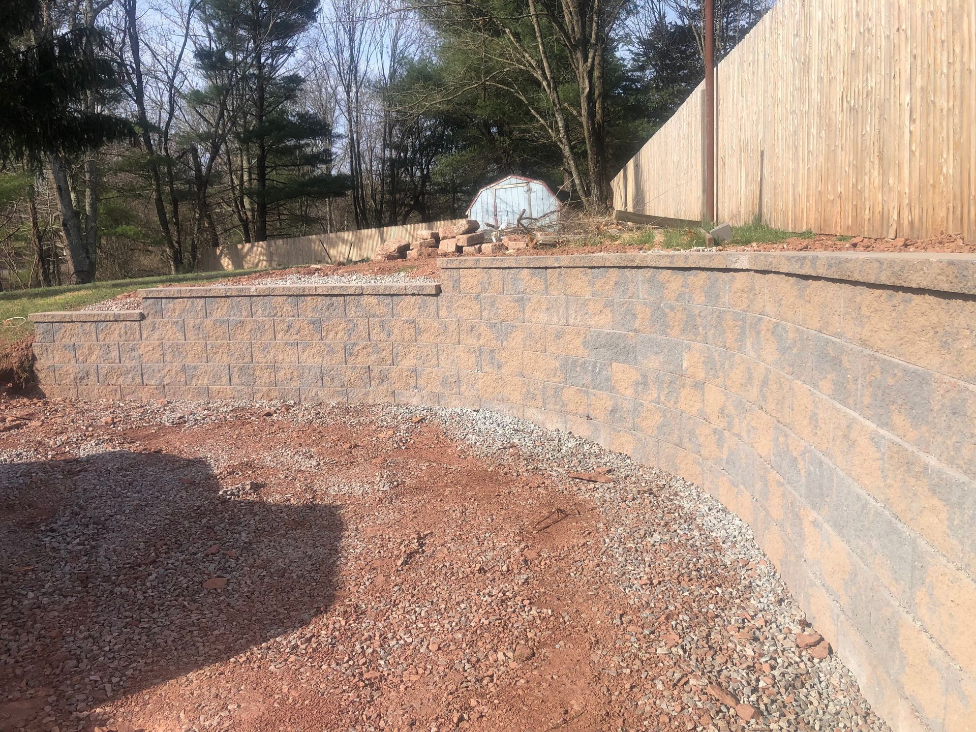 Curved stone retaining wall beside a gravel yard and wooden fence in a wooded backyard