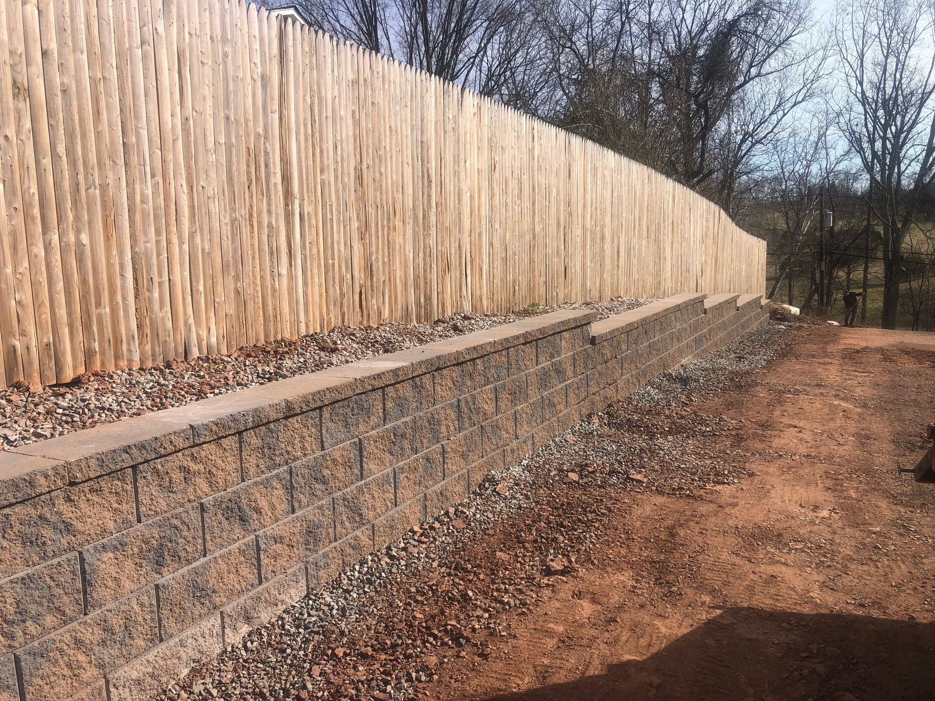 Wooden privacy fence atop a stone retaining wall along a dirt path in a wooded area