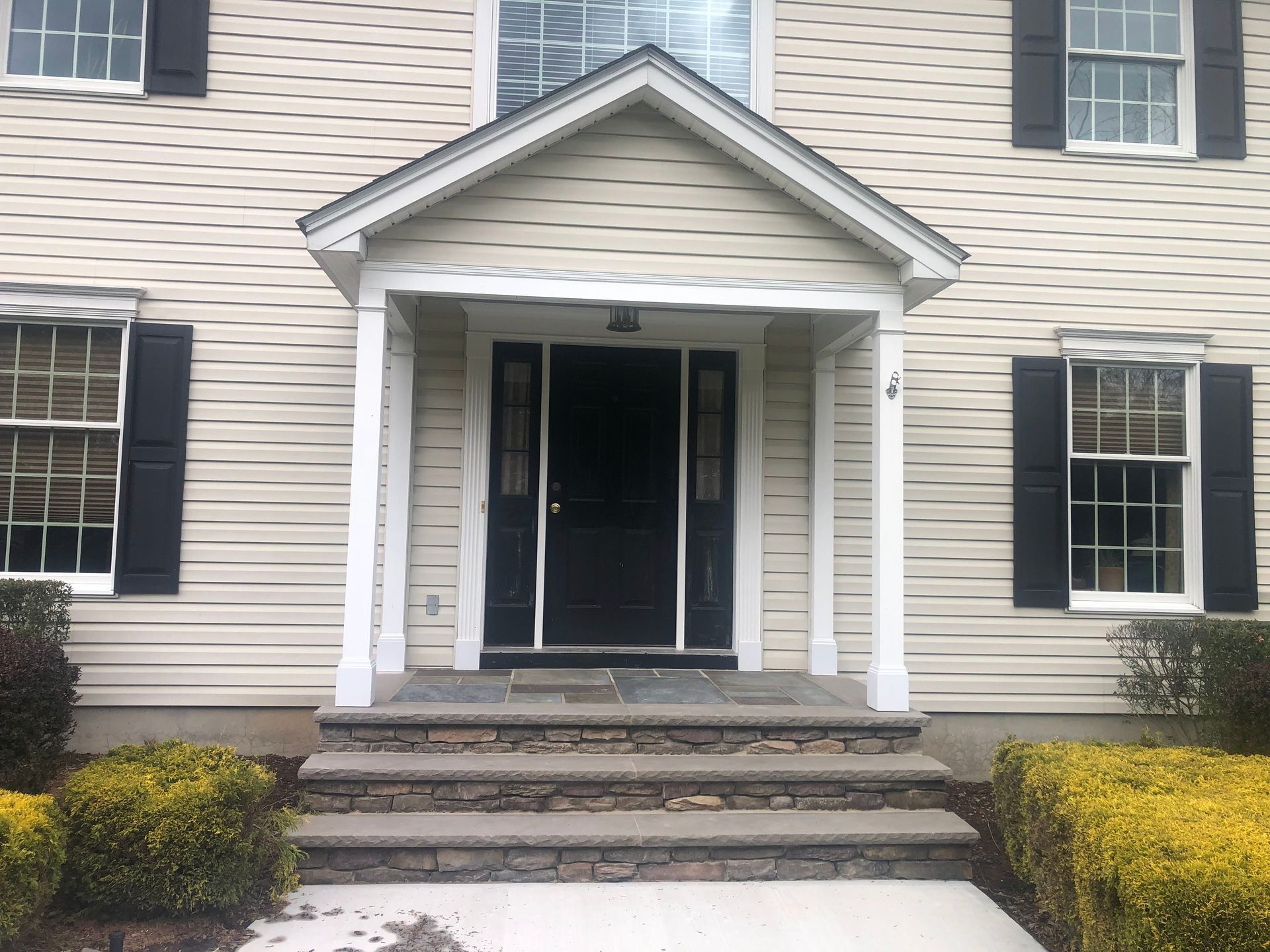 Front entrance of a beige house with white trim, black shutters, and stone steps leading to a covered porch