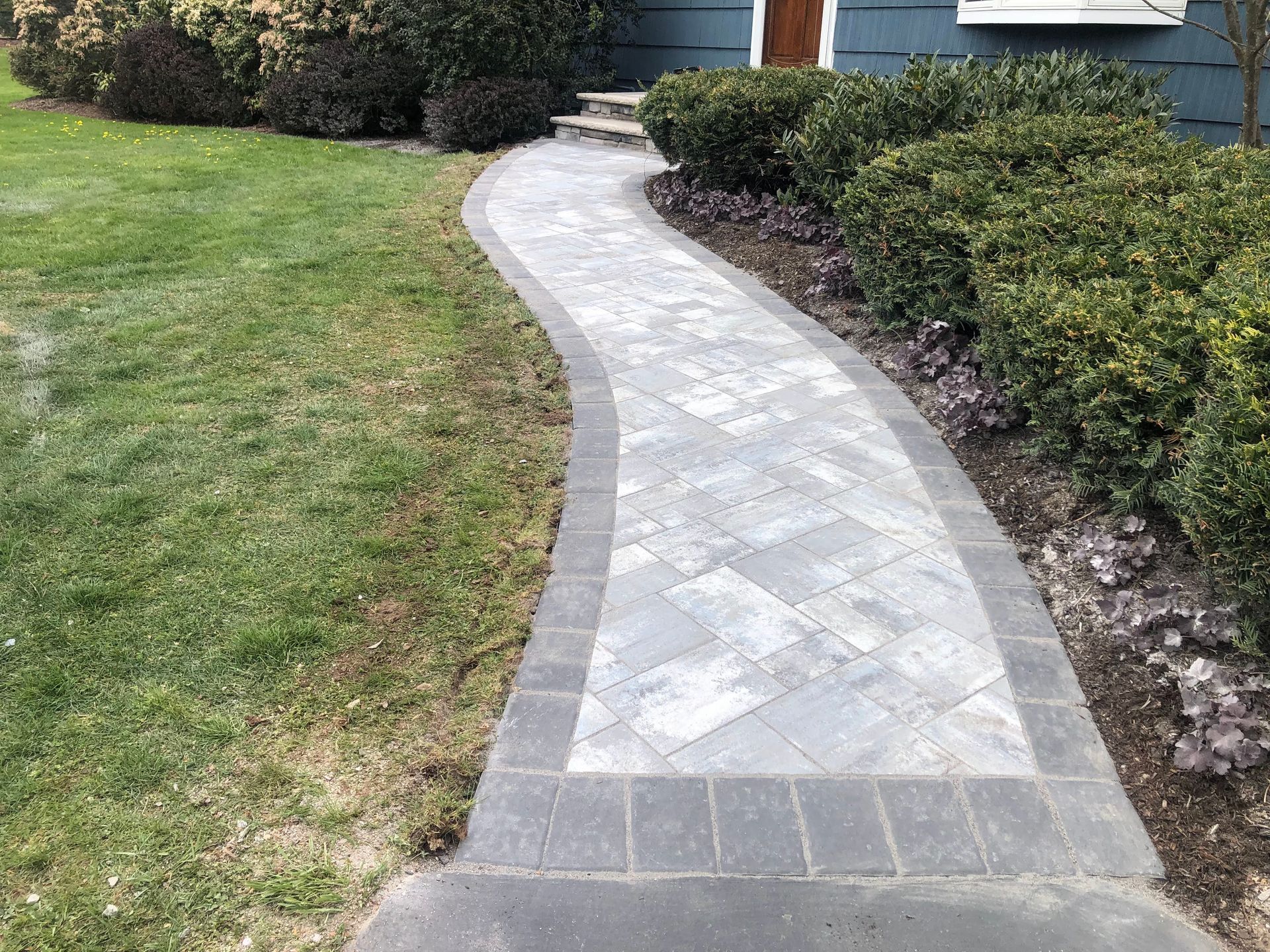 Curved gray stone walkway through a lawn beside trimmed hedges and a blue house