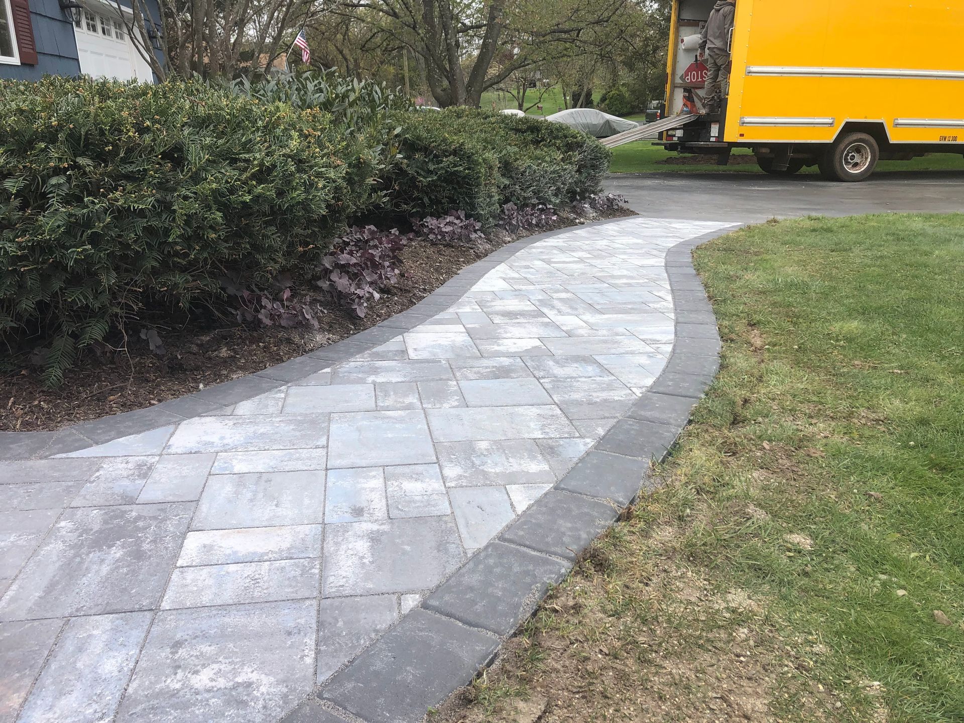 Curved stone walkway beside hedges and grass, with a yellow truck in the background.