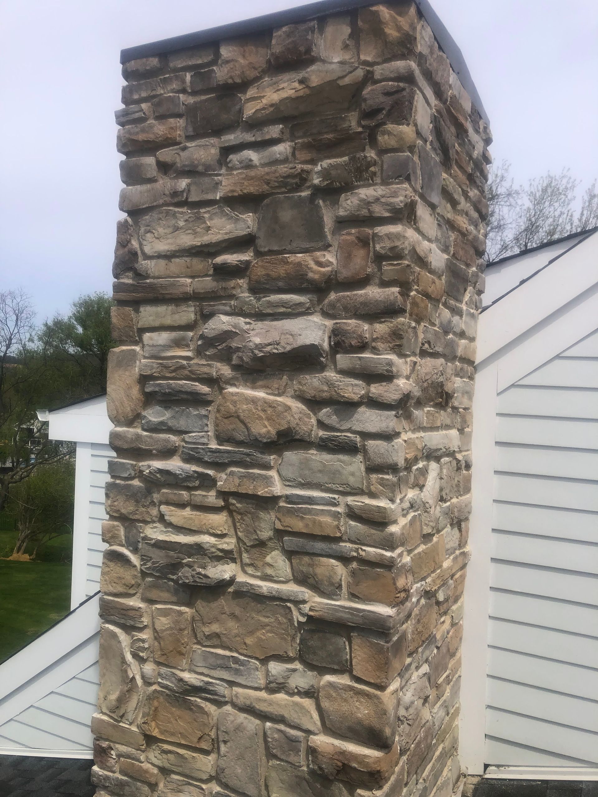 Stone chimney on the exterior of a house, with white siding and cloudy sky in the background