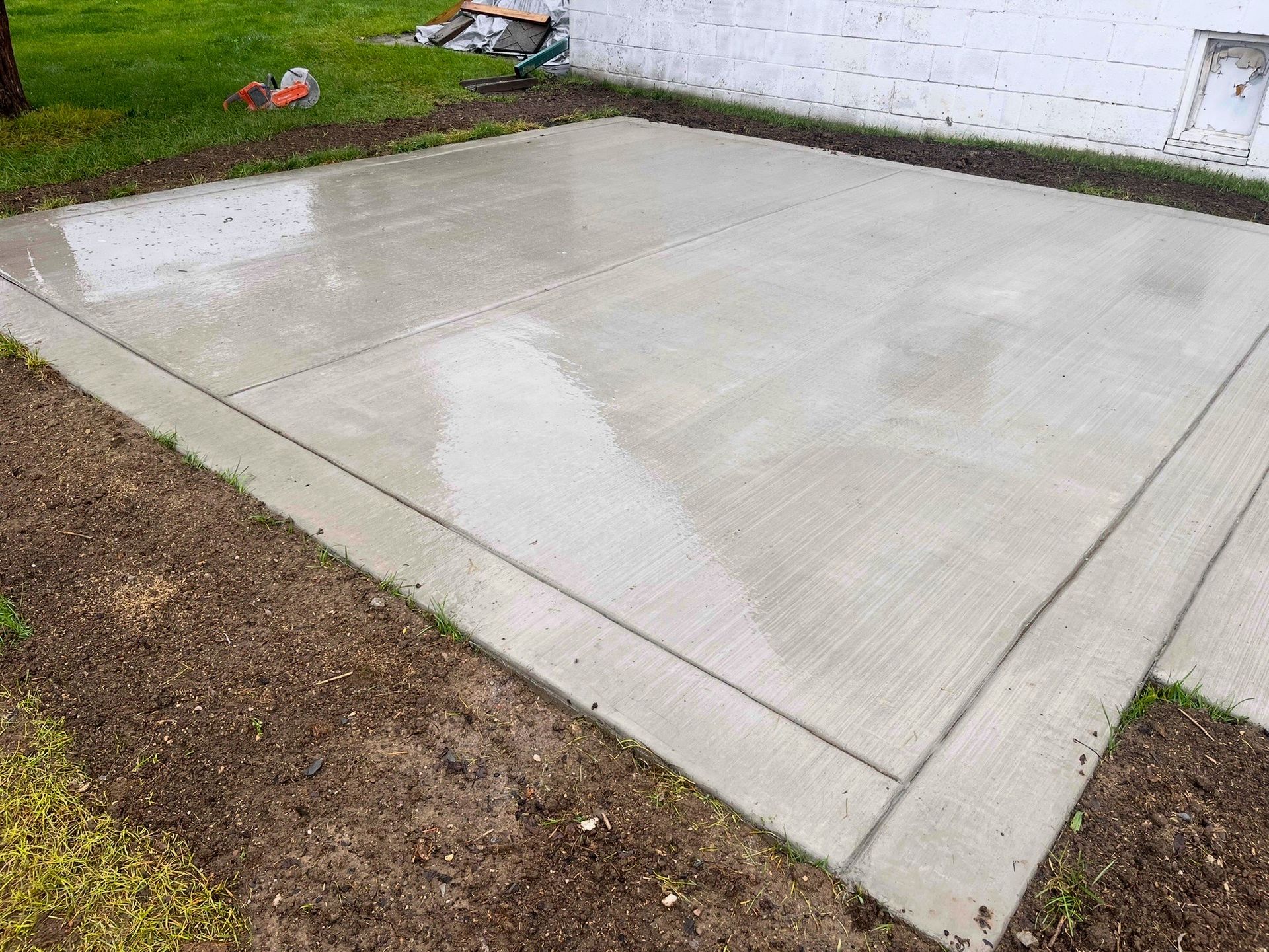 Freshly poured concrete patio beside a white wall and muddy yard, with wet surface and expansion joints.